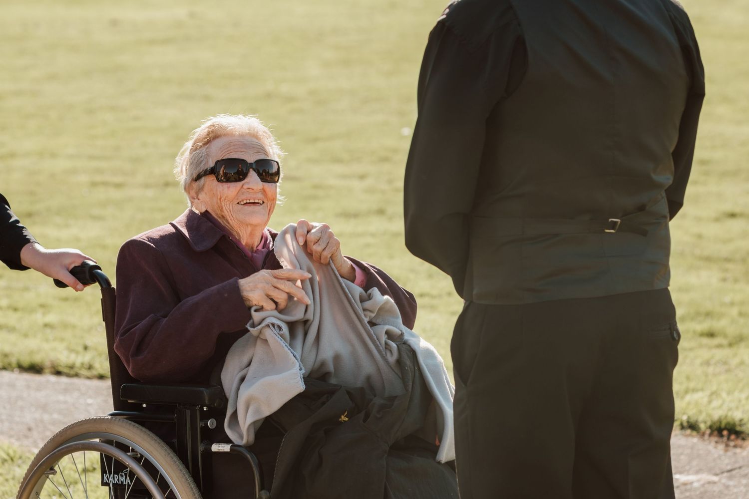 An elderly person in a wheelchair receives care assistance outdoors in natural sunlight on a grassy field.