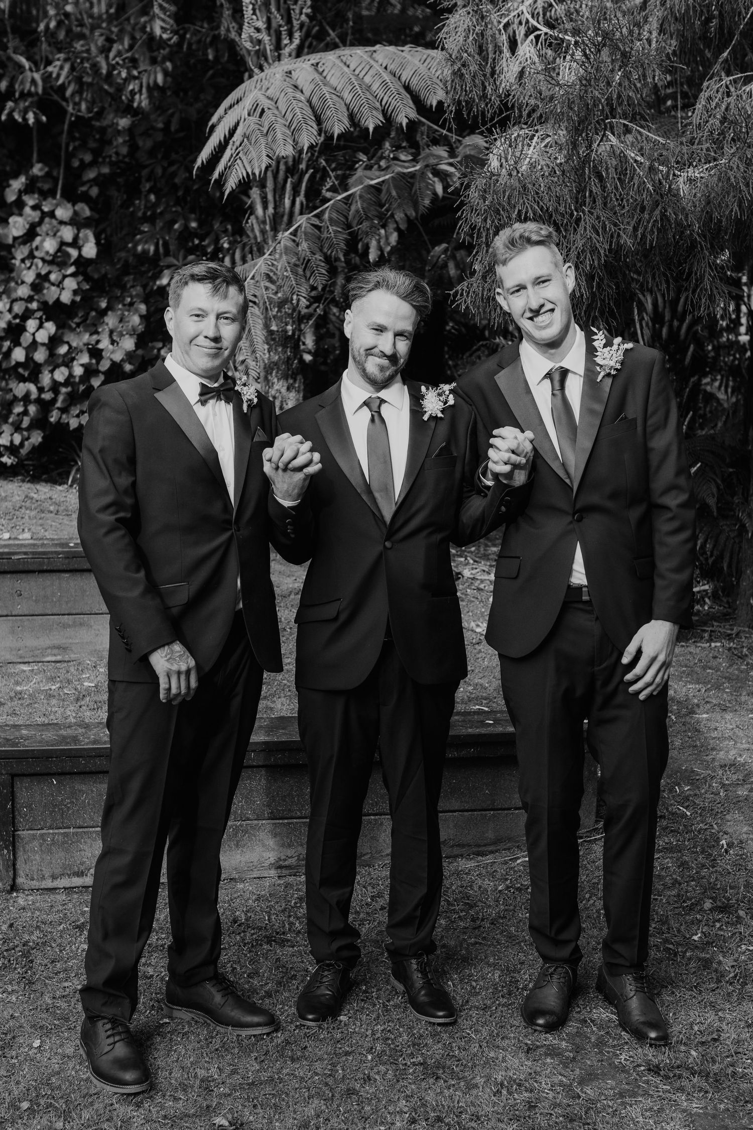 Three groomsmen in black suits pose playfully together for wedding photos outdoors in a garden setting.