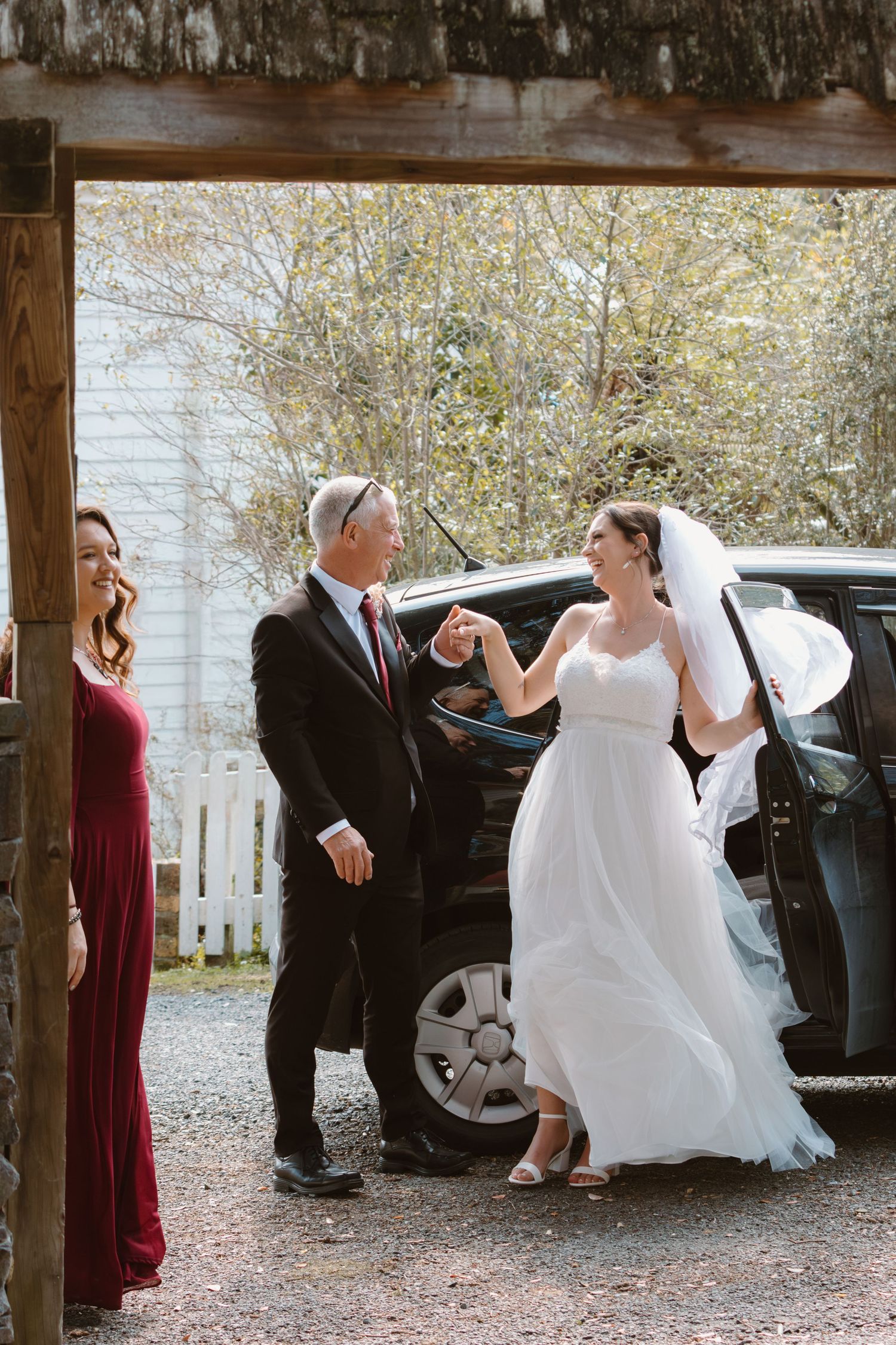 A wedding party arrives at a venue as a bride in a white gown steps out of a black car while someone in red looks on.