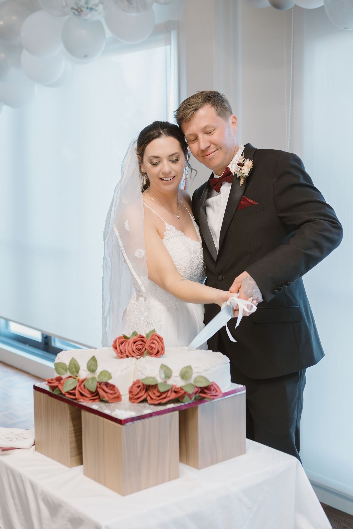 A bride and groom cut their elegant white wedding cake decorated with red roses and greenery at their reception.