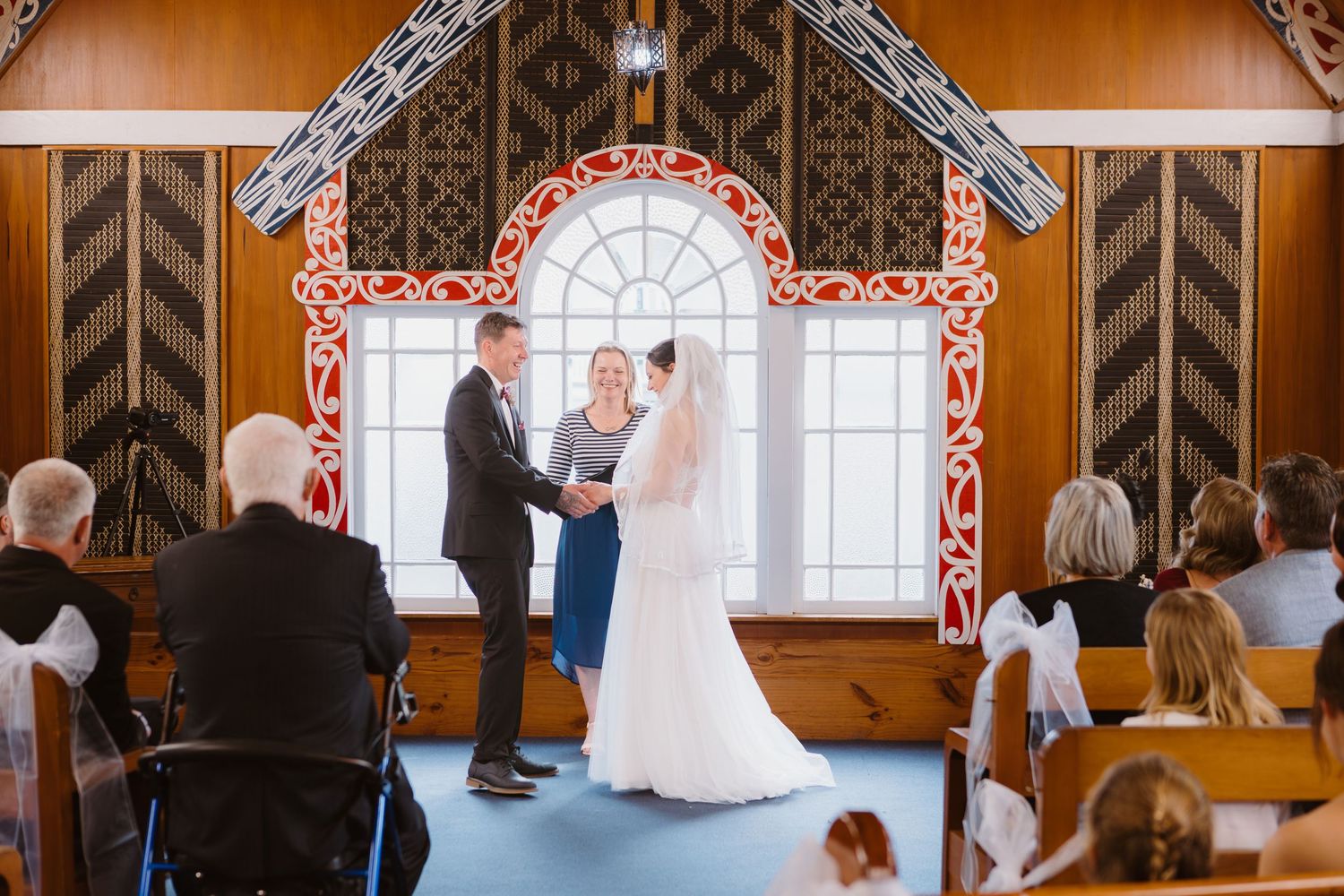 A wedding ceremony takes place inside a rustic wooden chapel with decorative window frames and wooden pews.