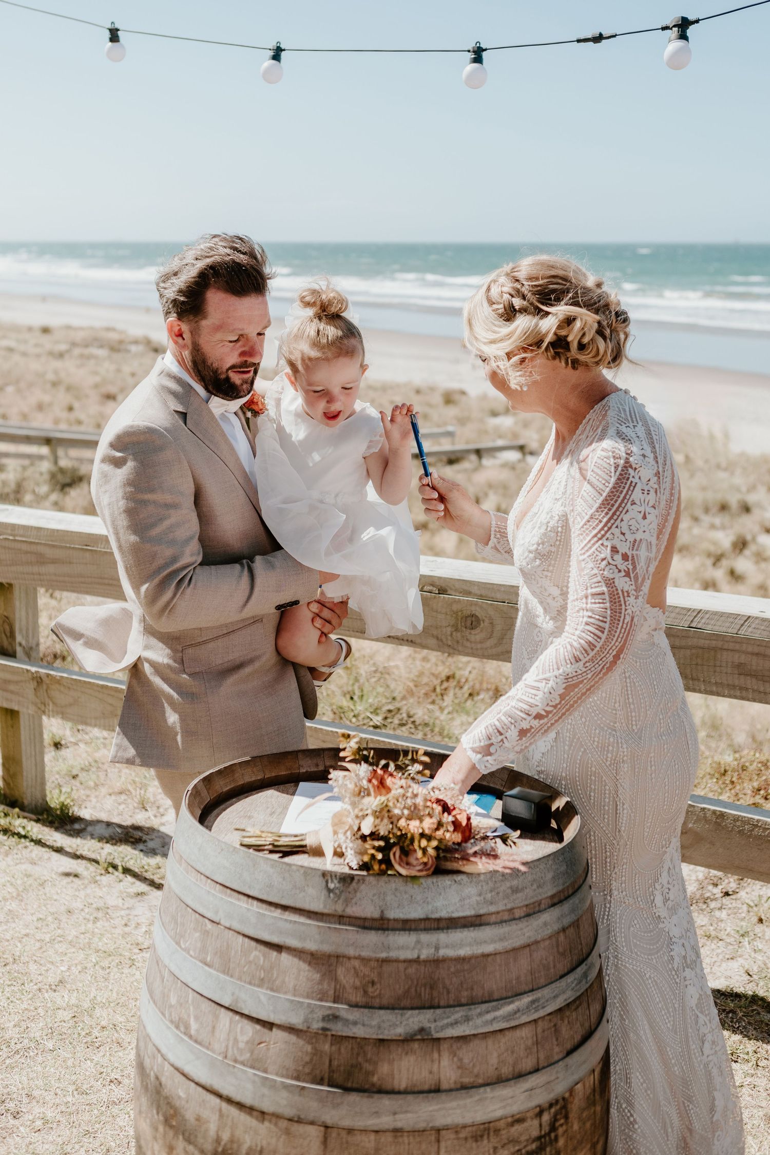 A beachside wedding ceremony takes place near a rustic barrel decorated with string lights and floral arrangements.