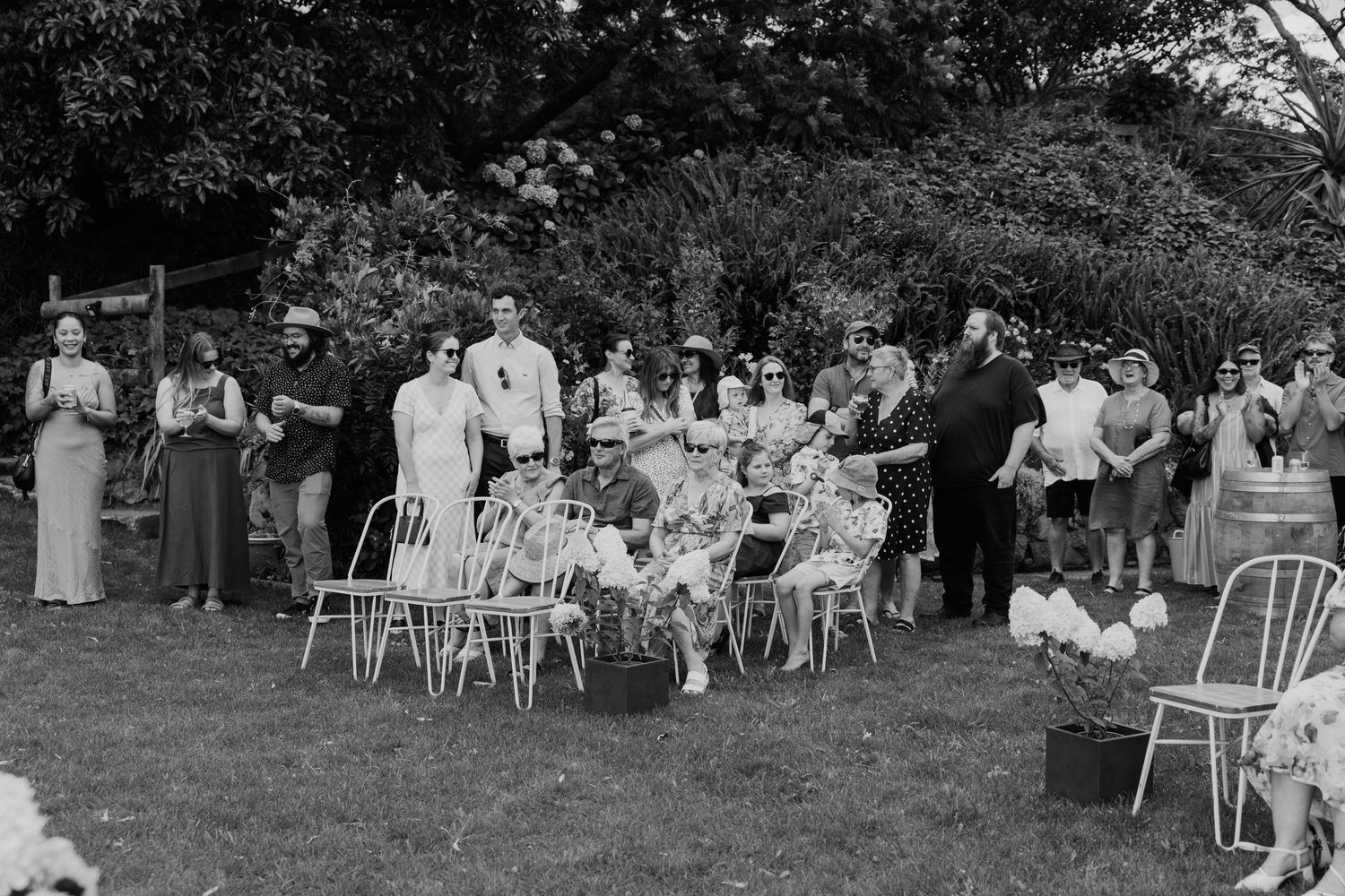 Black and white photo of guests gathered outdoors at a garden wedding ceremony, seated and standing on green lawn.