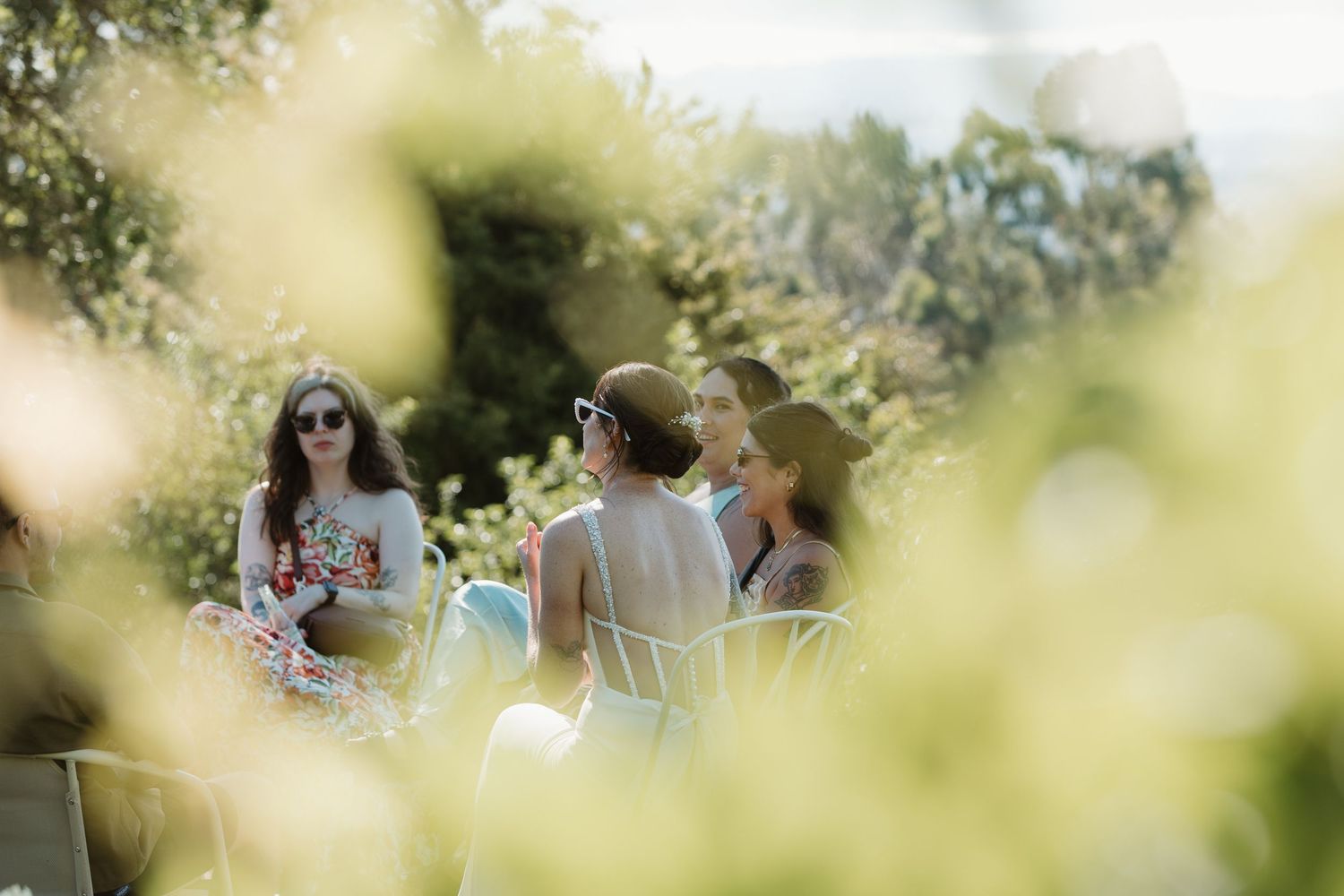 Women socializing outdoors in a sunlit garden, seated in white chairs surrounded by lush green foliage.
