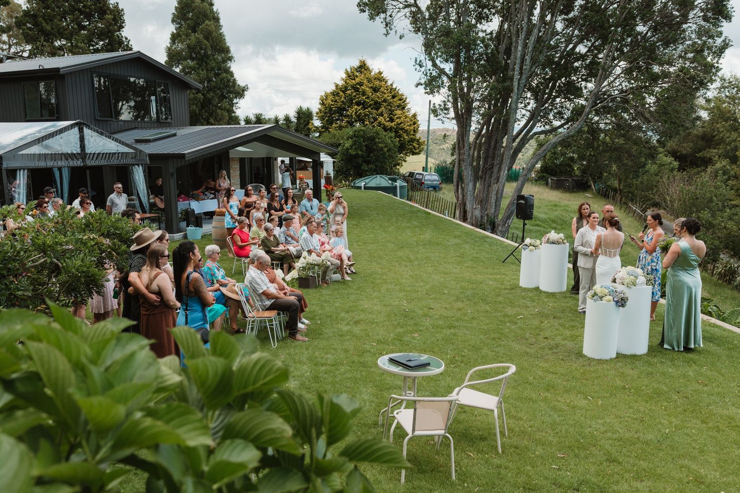 Outdoor garden wedding ceremony with guests seated on lawn beside elegant home surrounded by lush greenery.
