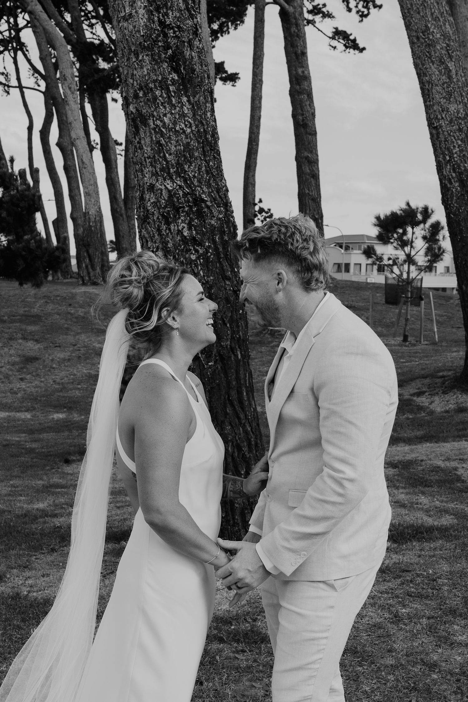 Black and white photo of a bride and groom holding hands, laughing together near trees on their wedding day.