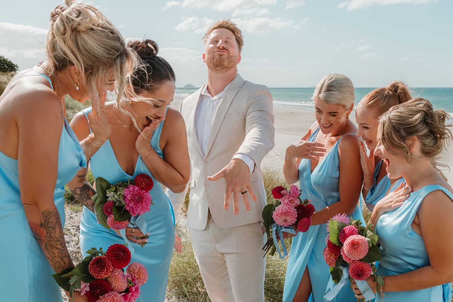 Groom in beige suit shows wedding ring to emotional bridesmaids in blue dresses holding pink bouquets on a beach.