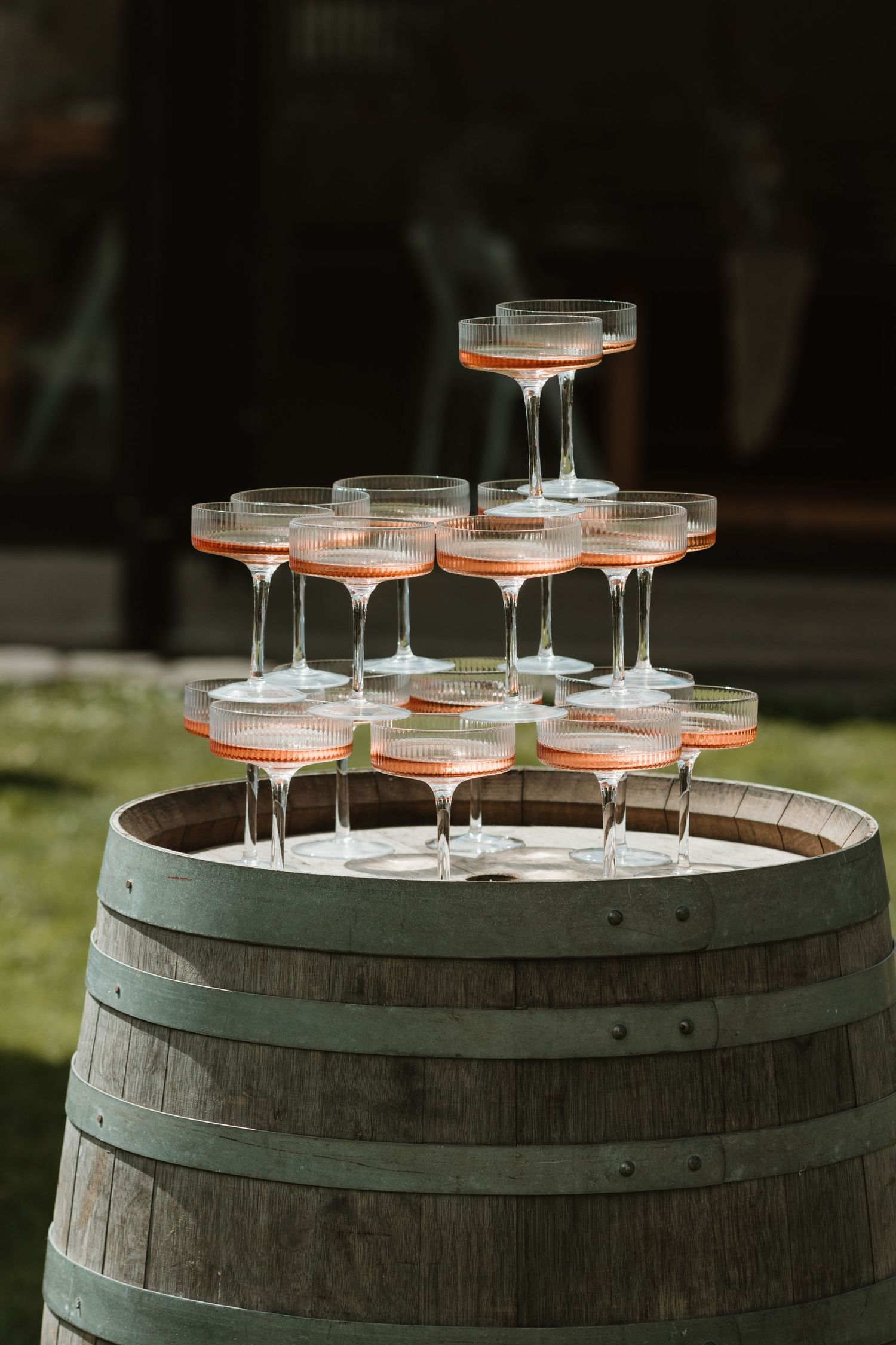 Champagne coupe glasses arranged in a pyramid stack on top of a rustic wooden wine barrel outdoors.