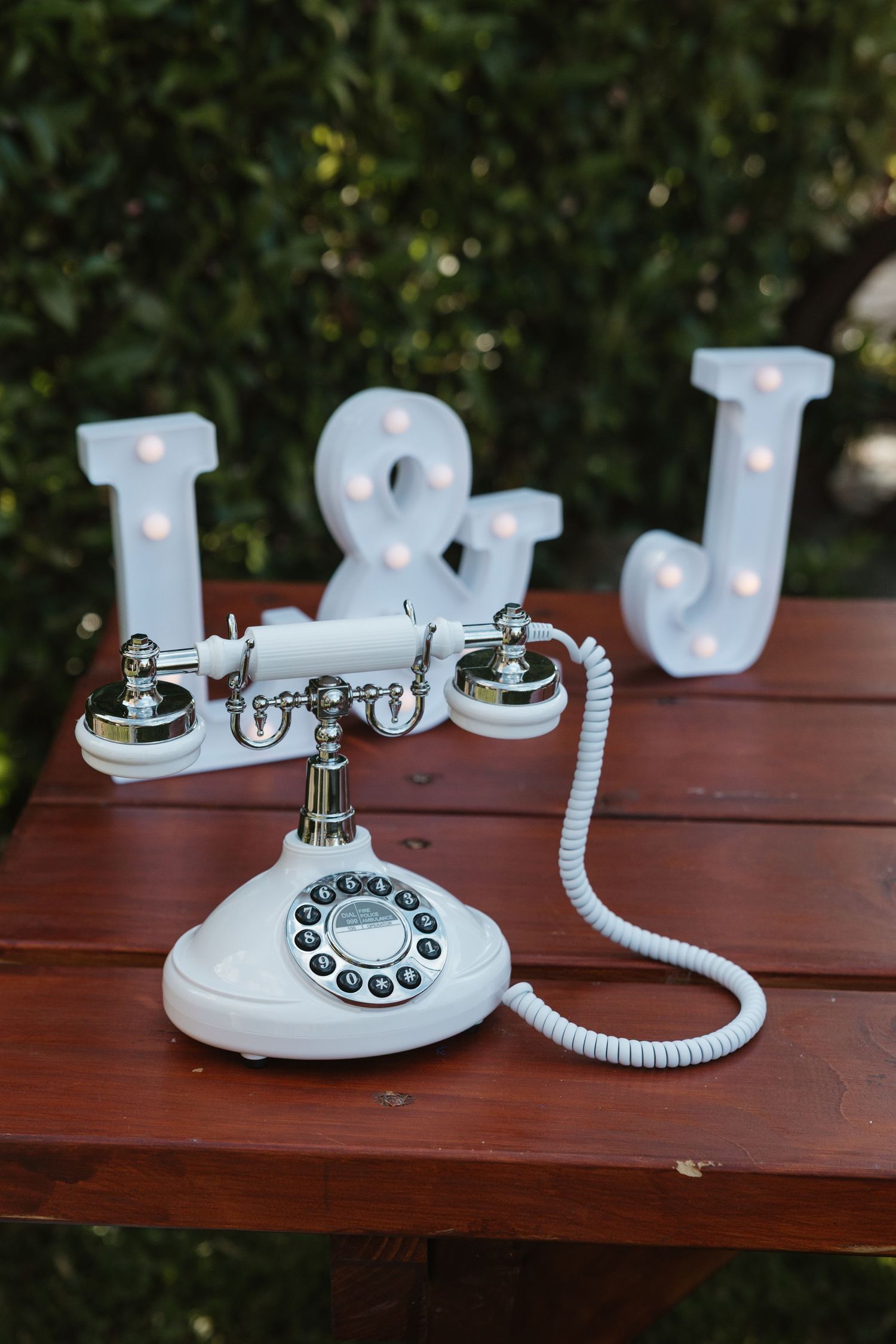 A vintage-style white rotary telephone displayed on a wooden outdoor table with decorative LED letters in the background.