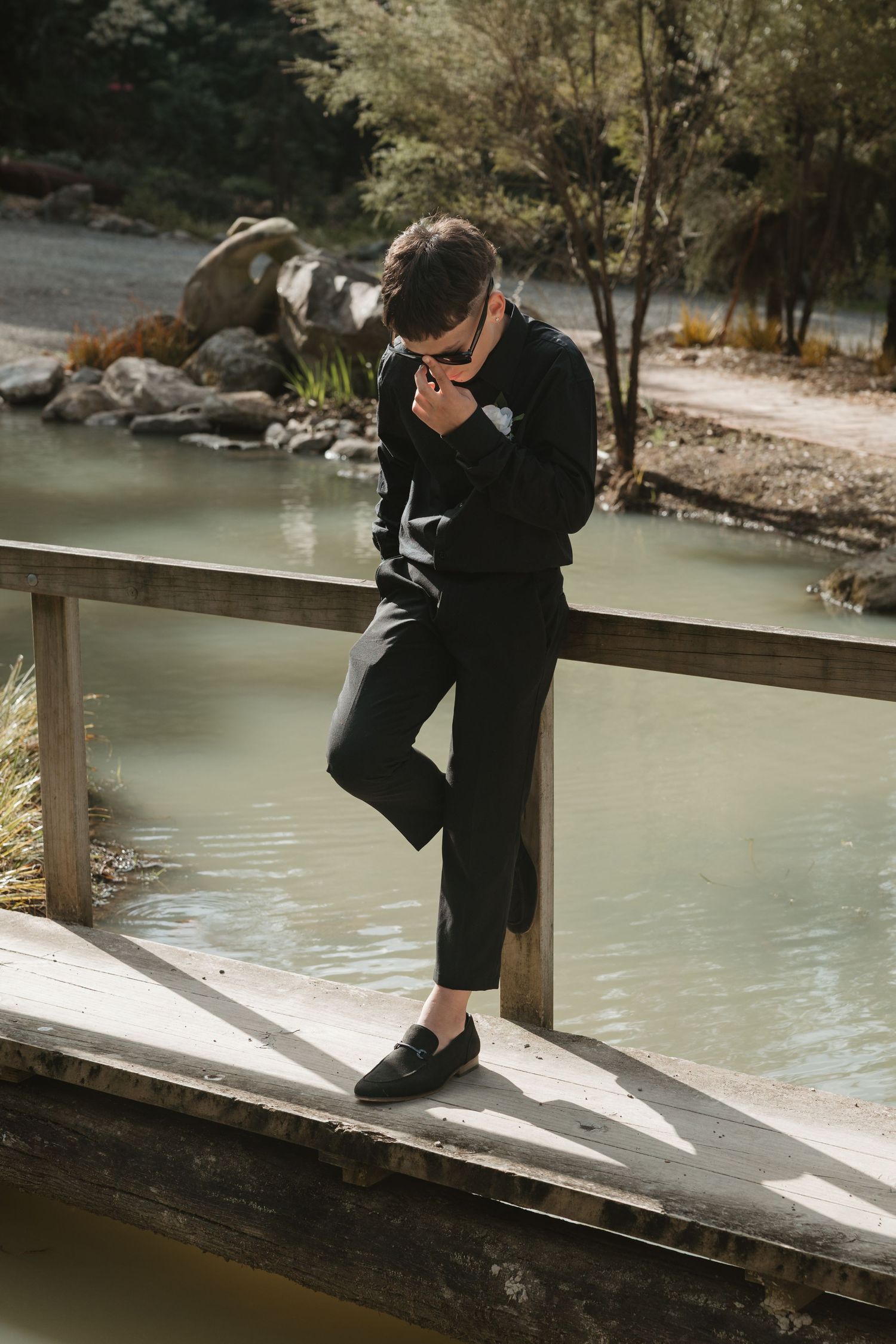 Person in dark clothing sits on wooden railing by calm river in natural outdoor setting.