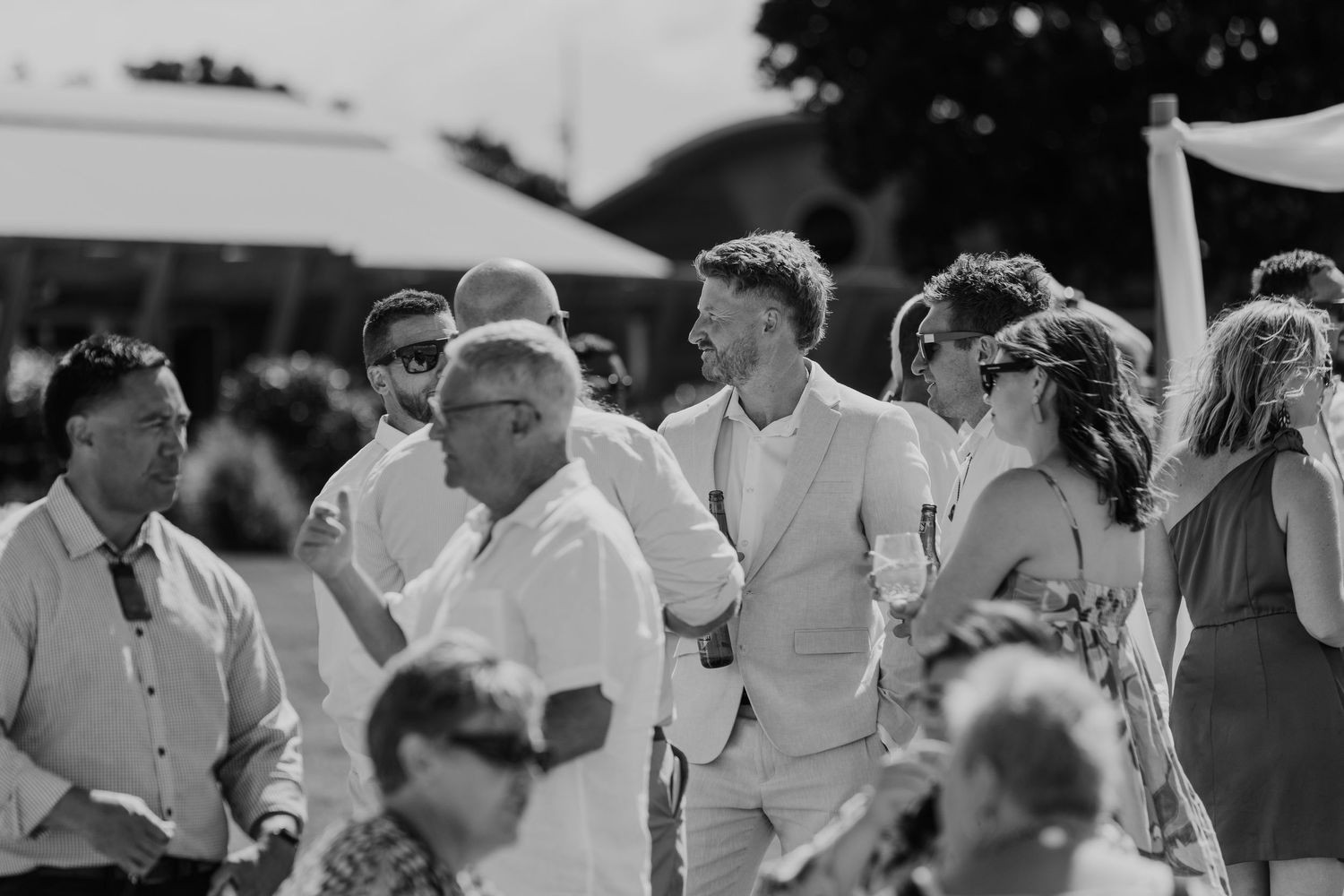 Black and white photo of elegantly dressed wedding guests socializing outdoors at a sunny reception.