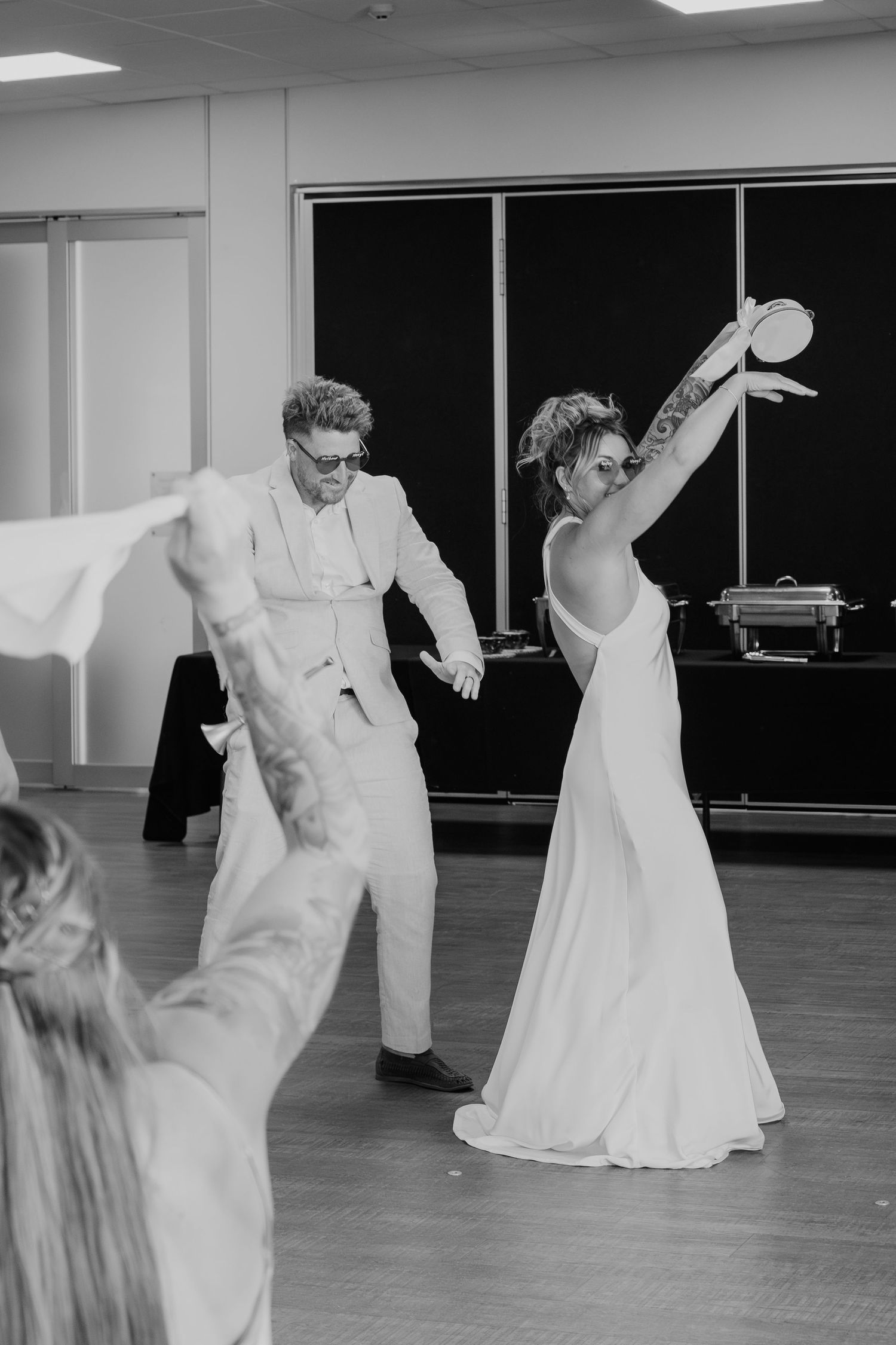Black and white photo of a bride and groom dancing joyfully at their wedding reception in a bright venue.