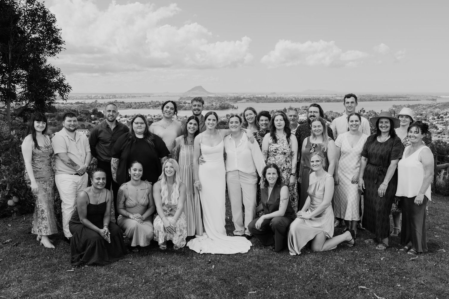 Black and white wedding group photo outdoors with bride and guests posing together on a scenic hillside.