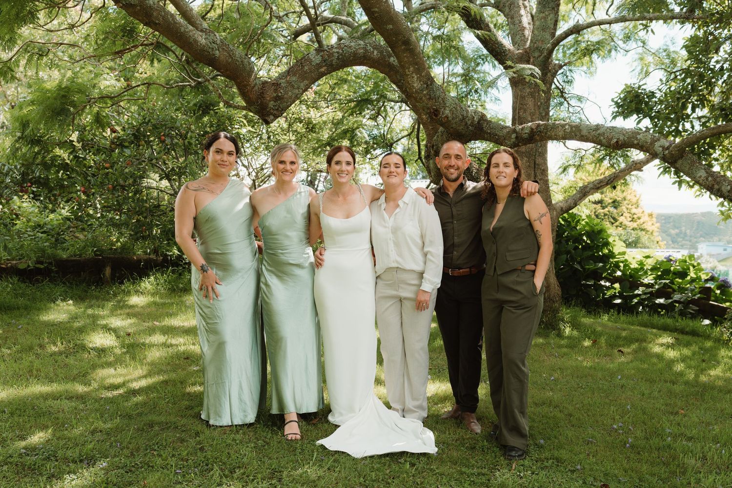 Wedding party poses outdoors under large tree, bride in white gown with bridesmaids in sage green dresses.