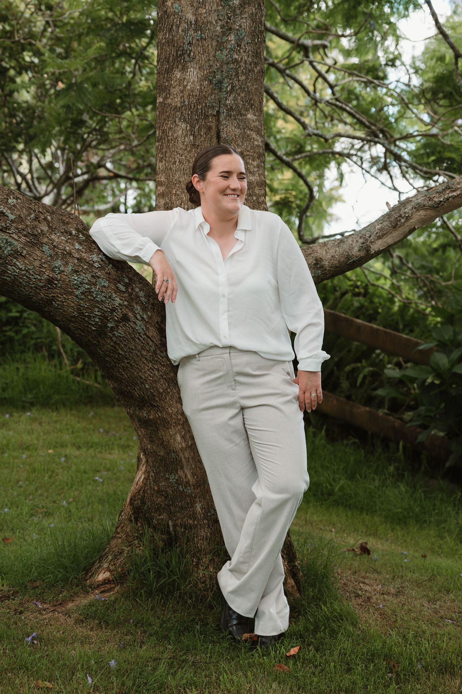 Person in white shirt and light pants leaning against a tree in a lush green outdoor setting.
