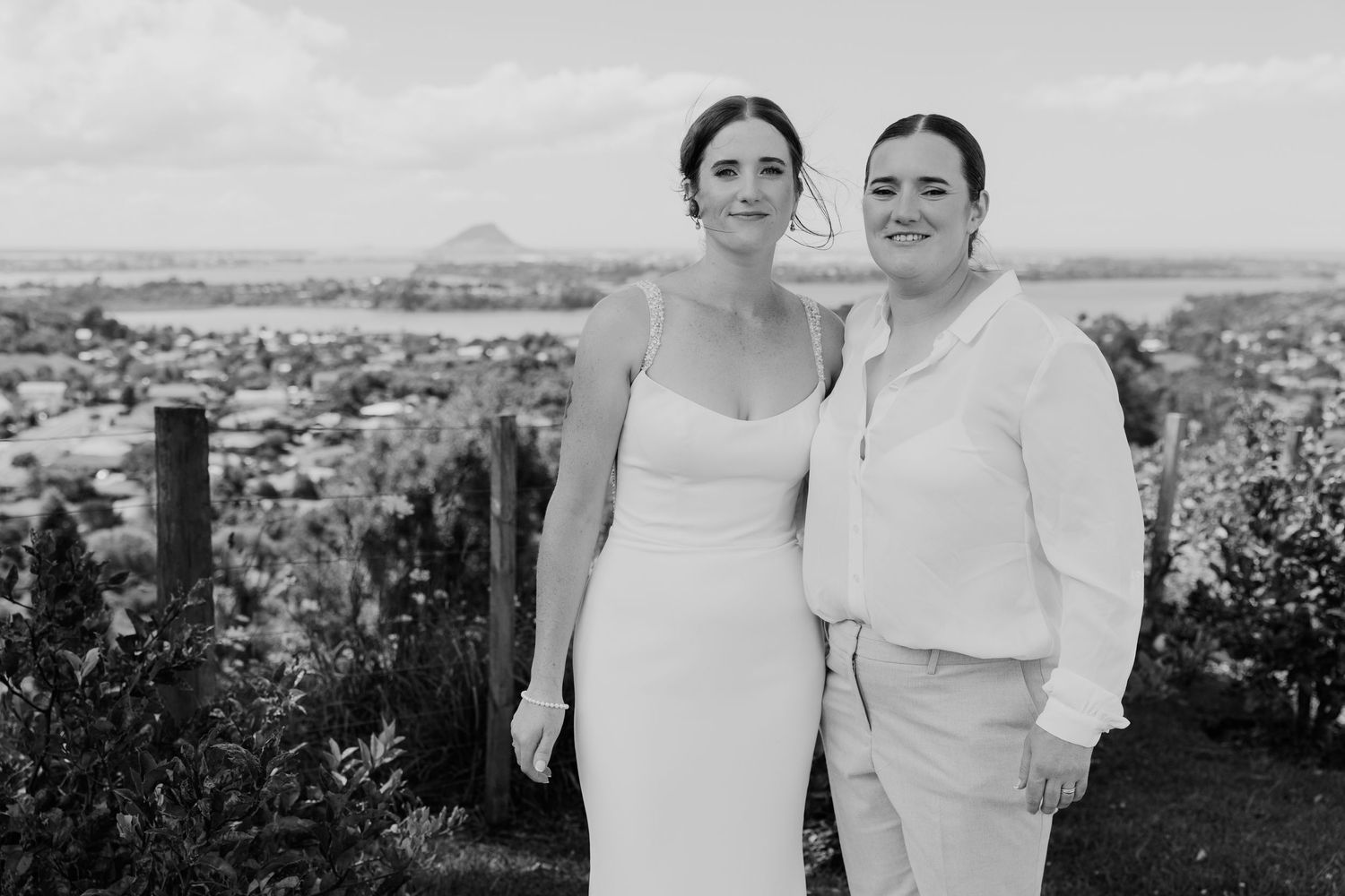 Two brides pose together smiling on a hilltop with a scenic coastal town and mountain backdrop on their wedding day.