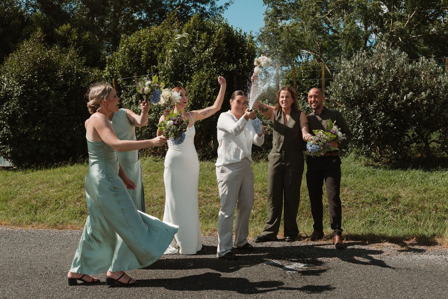 Wedding party celebrating outdoors, bride in white gown with bridesmaids in sage green dresses and groomsmen holding bouquets.