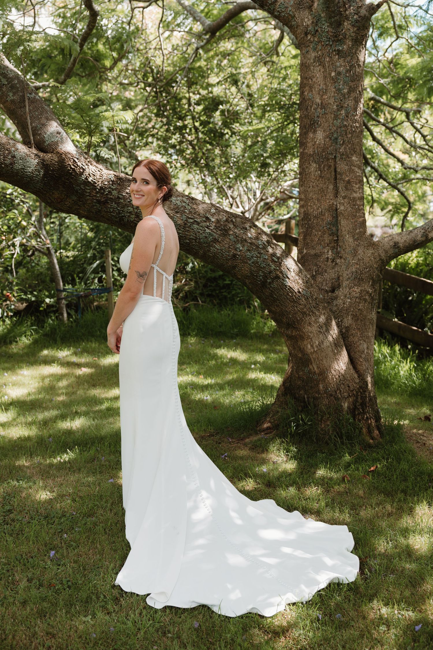 Bride in elegant white backless gown with train stands by large tree in lush garden, smiling over shoulder.