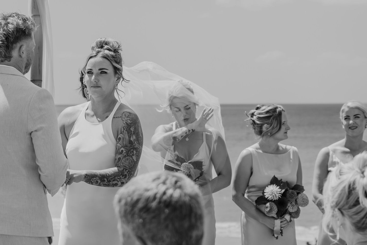 Black and white beach wedding ceremony with tattooed bride in veil, emotional bridesmaid wiping tears, ocean backdrop.