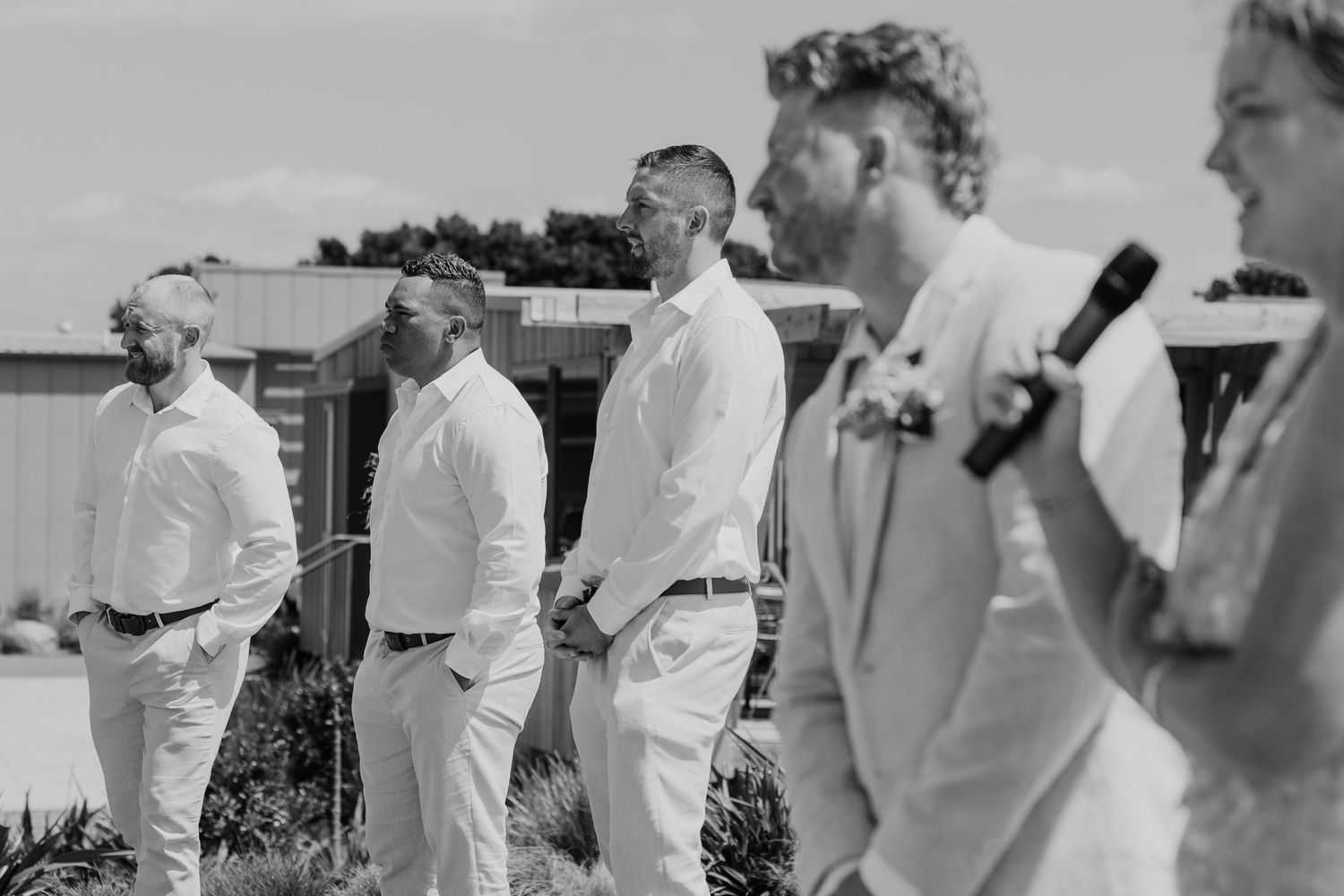 Black and white photo of groomsmen standing outdoors at a wedding ceremony, listening attentively to a speech.