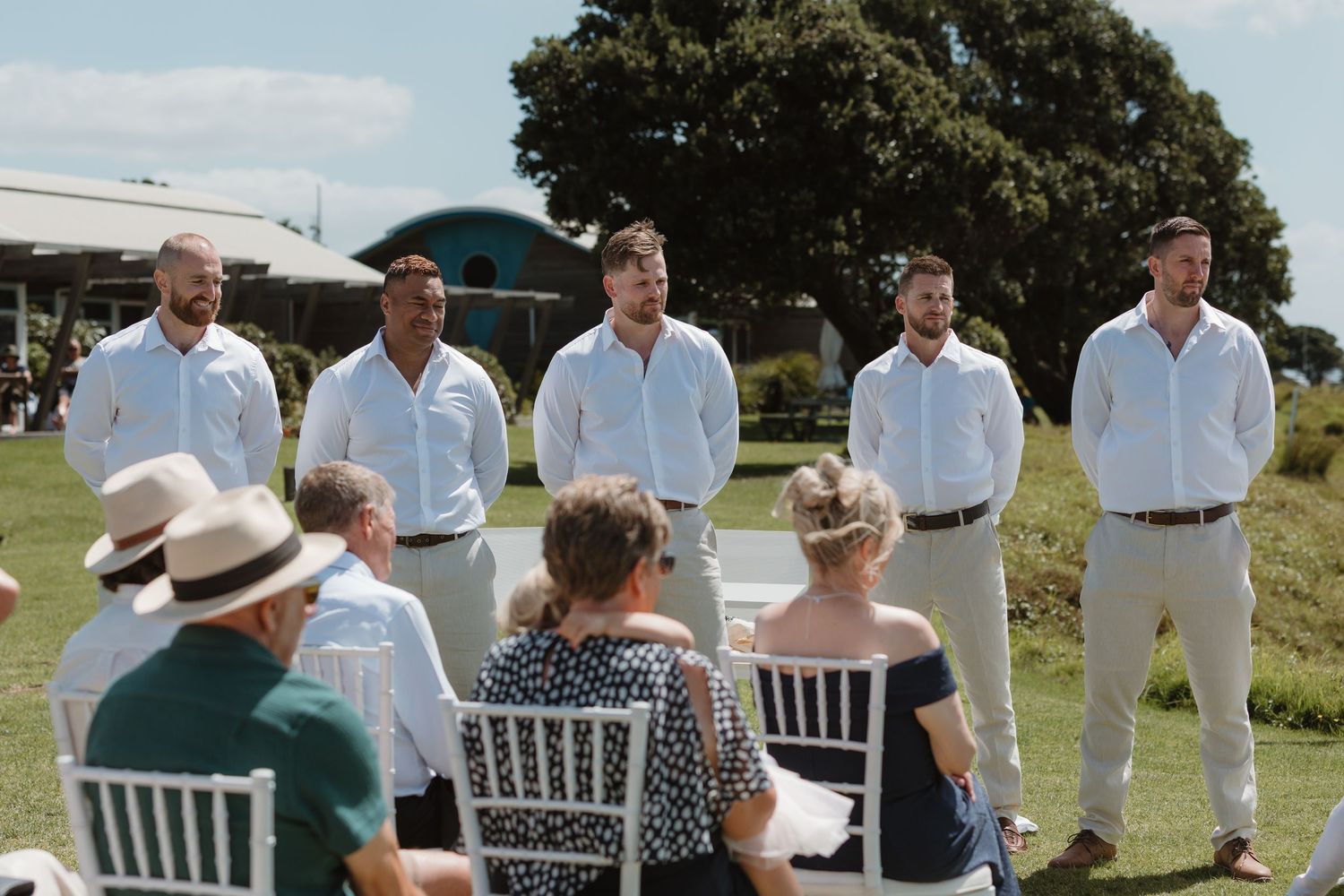 Groomsmen in white shirts stand at outdoor wedding ceremony while guests sit in white chairs on green lawn.