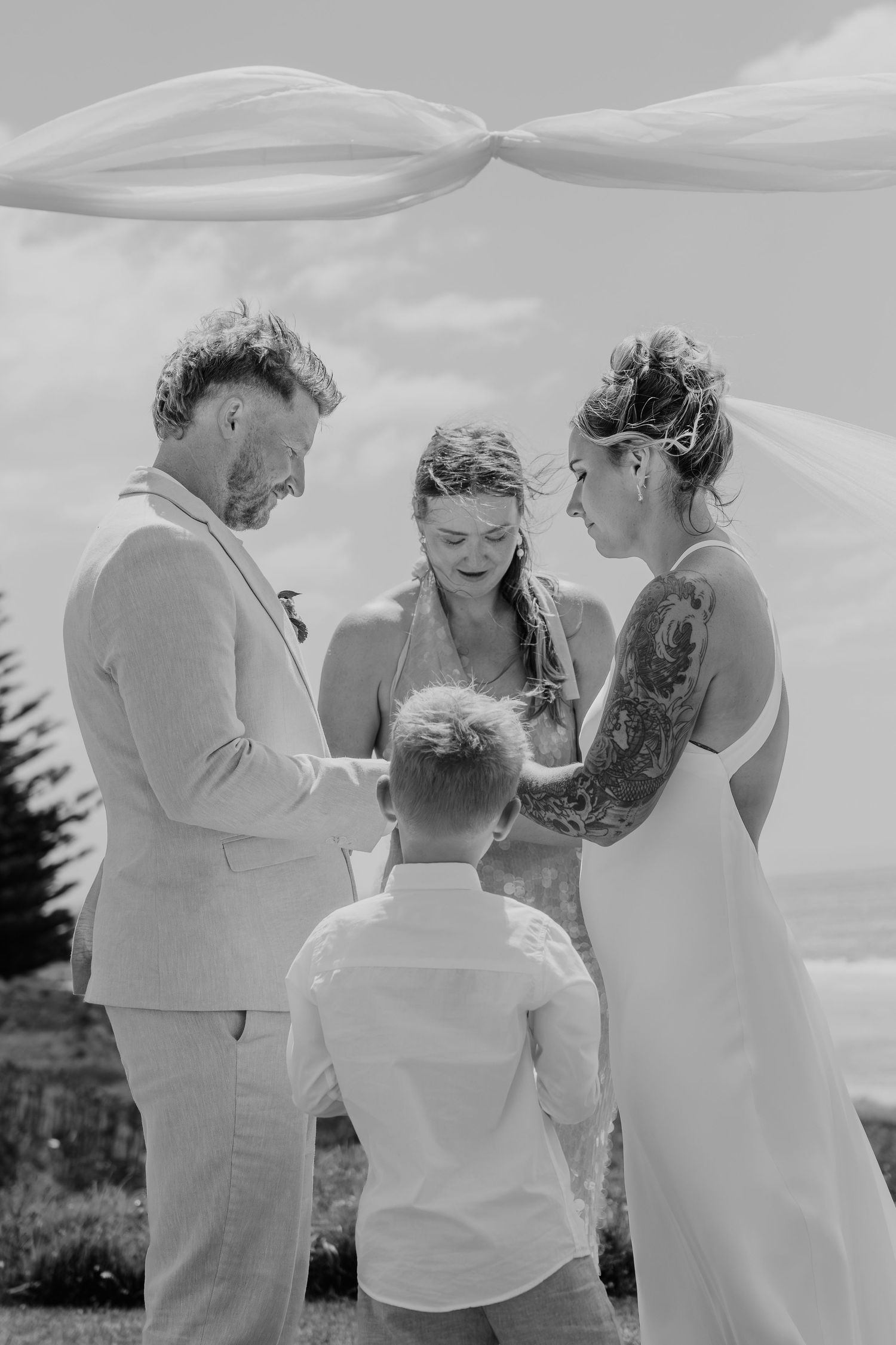 Black and white wedding ceremony photo with couple, officiant, and child holding hands outdoors under flowing fabric.