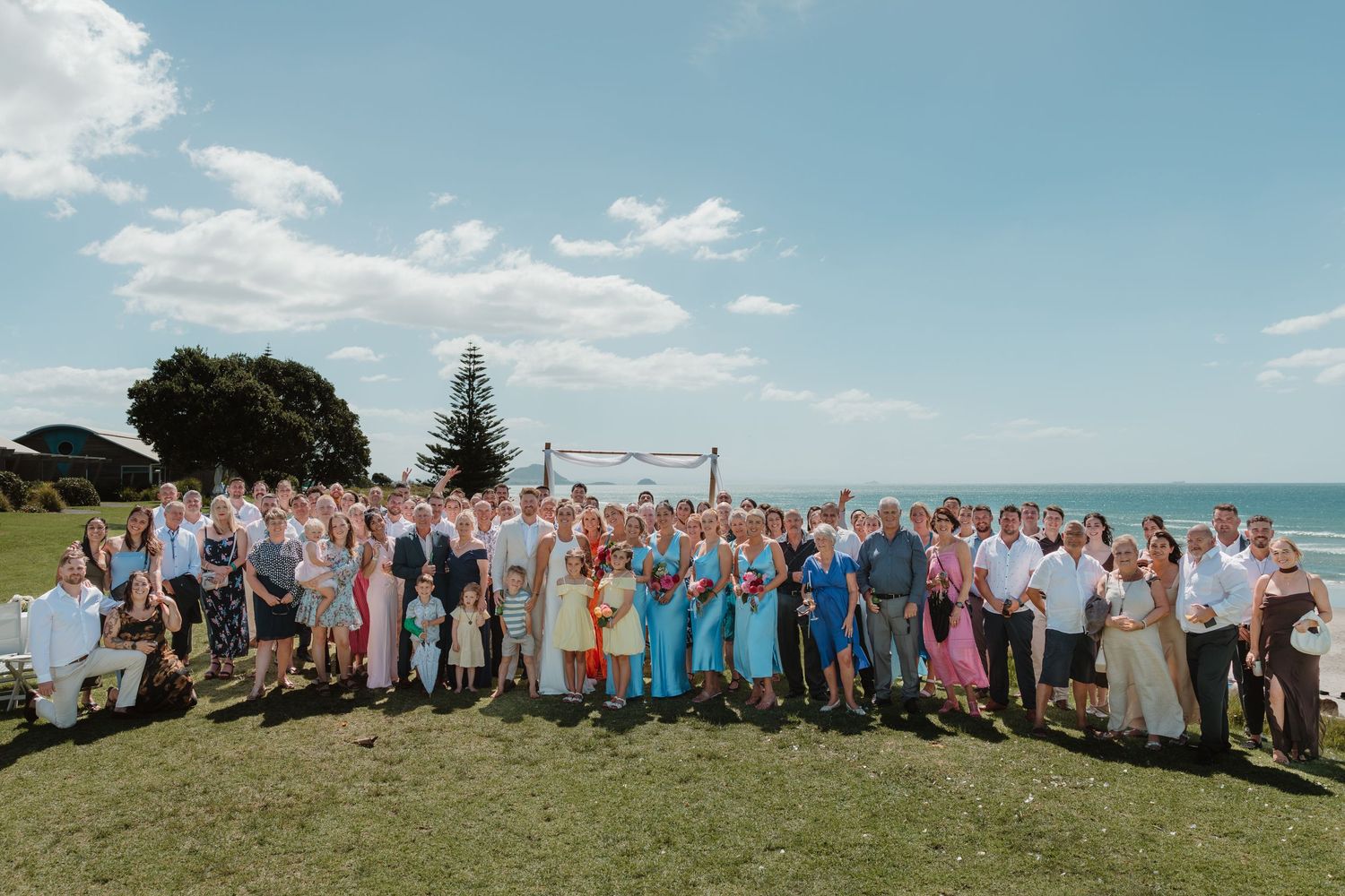 Large wedding group photo outdoors by the ocean on a sunny day with blue skies and lush green grass.