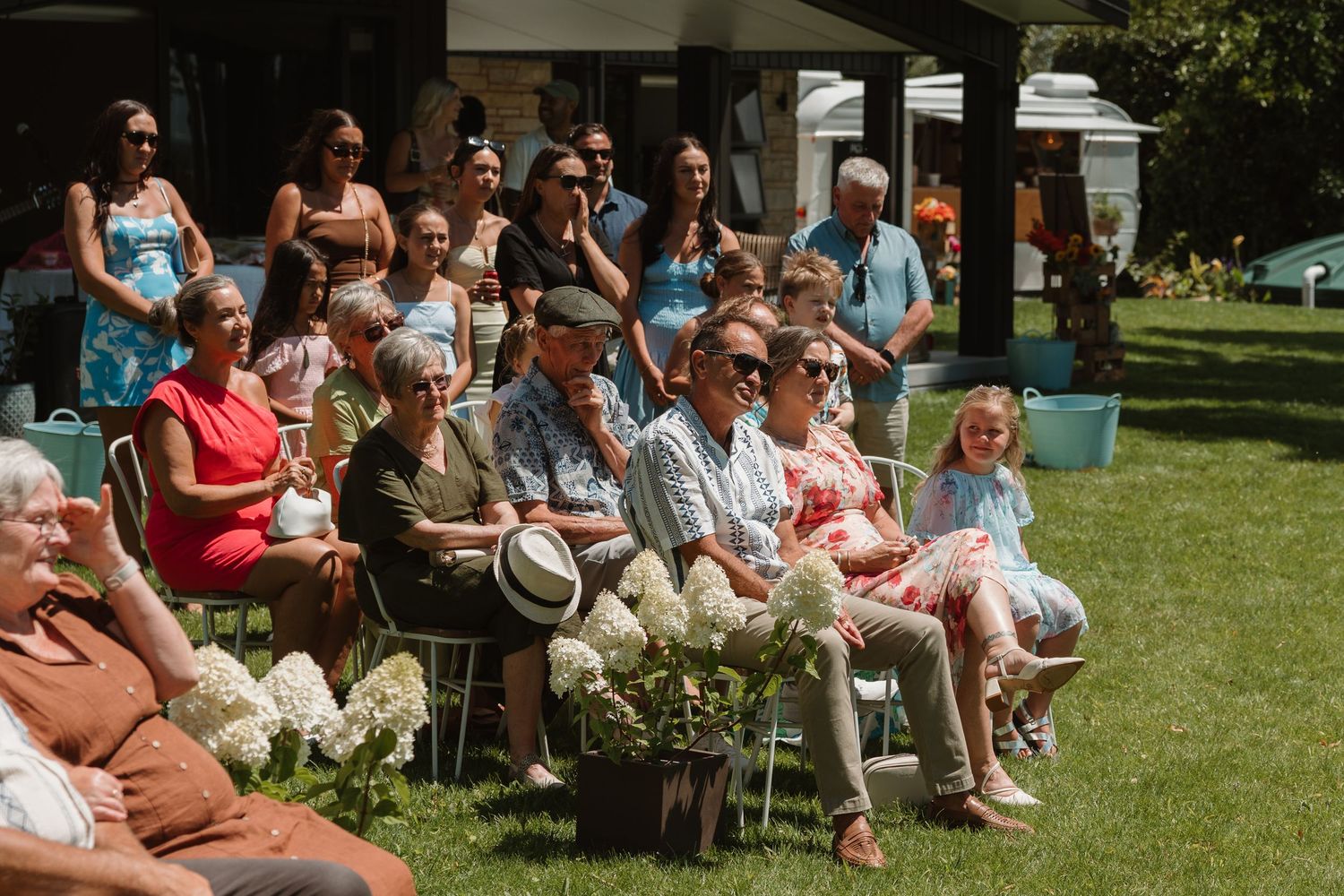 Guests seated outdoors on lawn chairs at a sunny garden wedding ceremony, surrounded by white hydrangeas.
