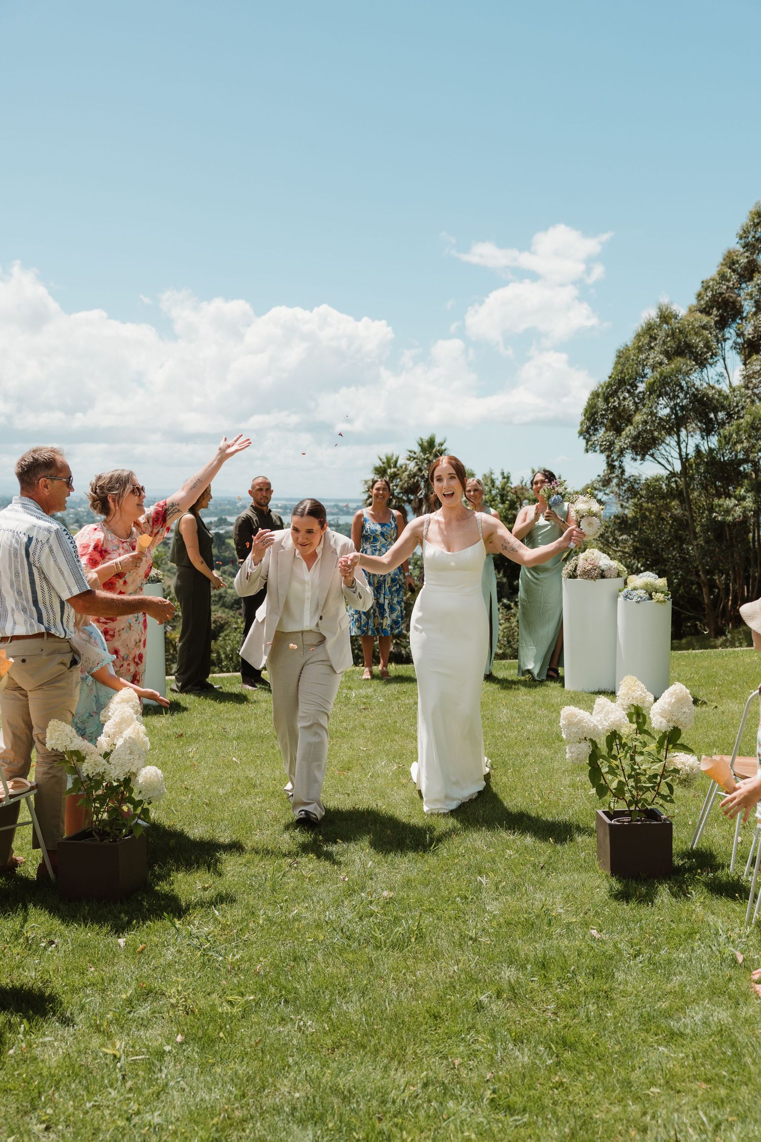 Two brides walk joyfully down outdoor aisle as guests celebrate their sunny garden wedding ceremony.