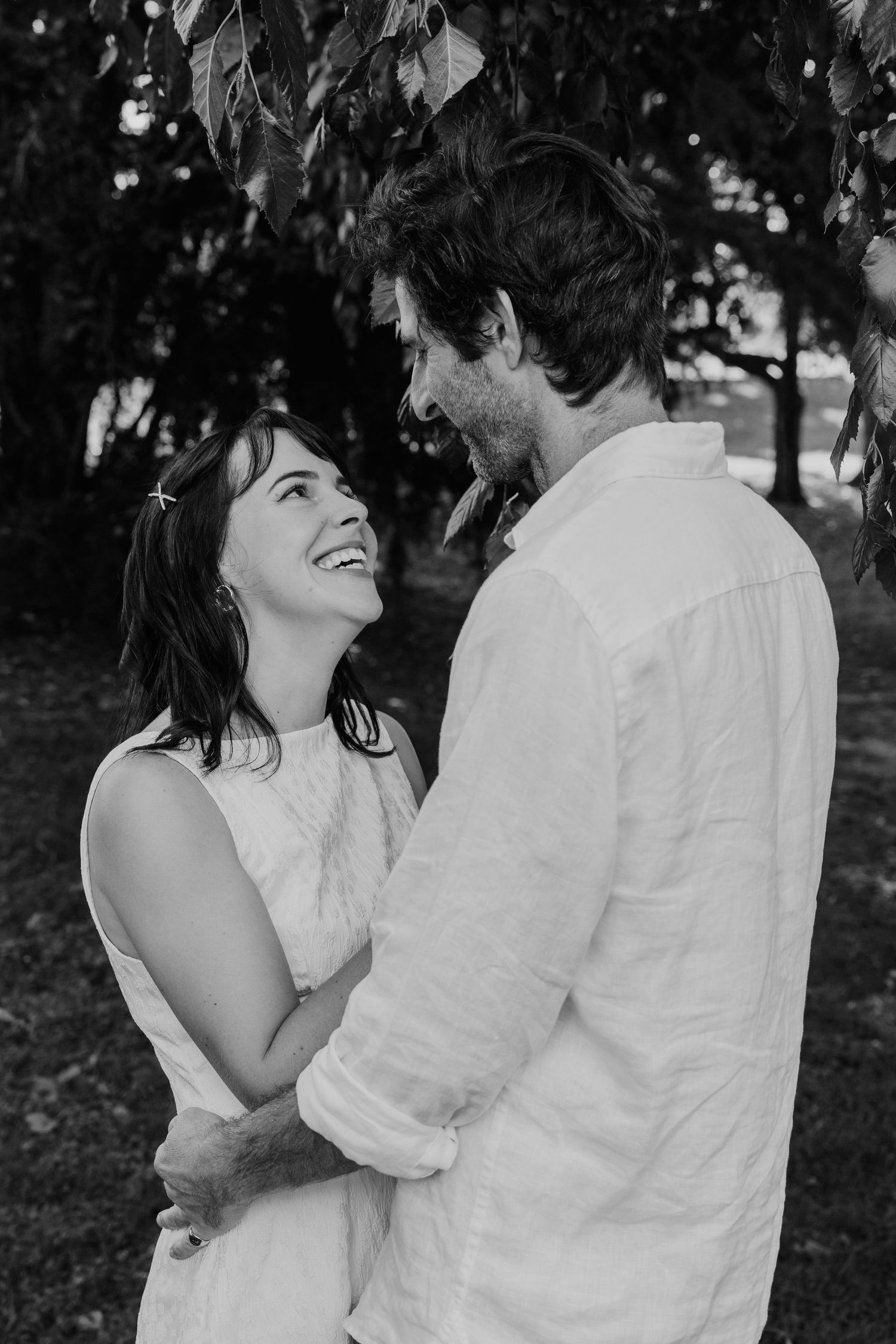 Black and white photo of a laughing couple embracing outdoors under leafy trees, wearing white outfits.