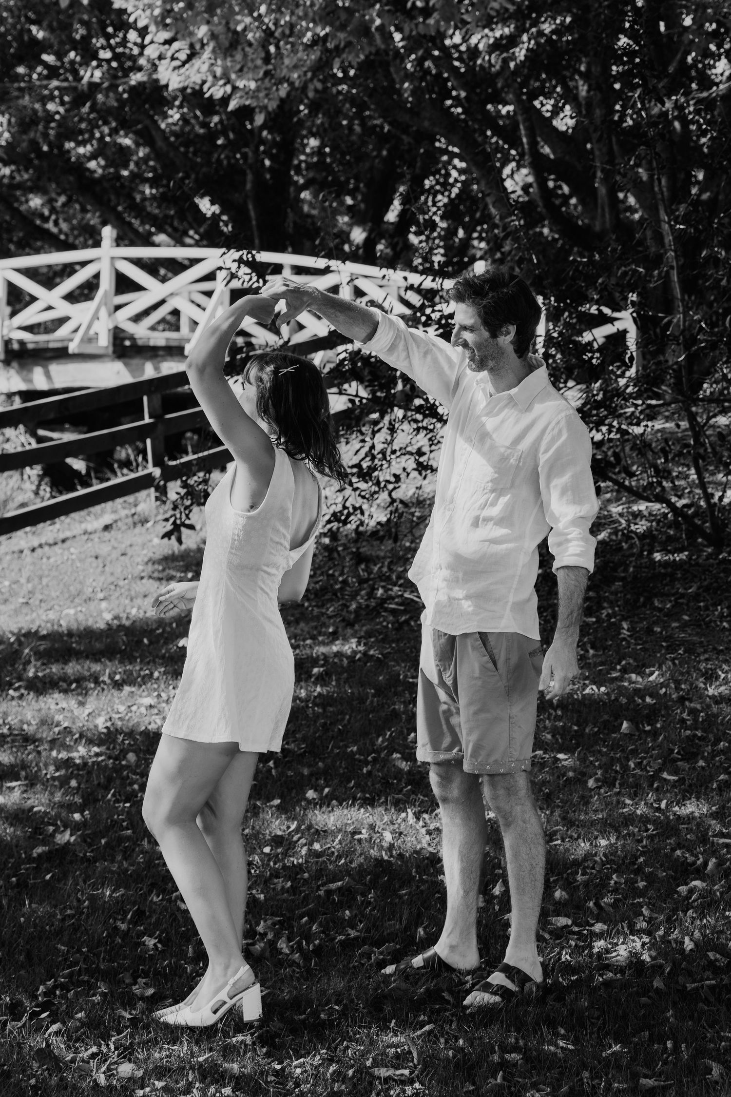 Black and white photo of a couple dancing outdoors near a white bridge surrounded by lush trees.