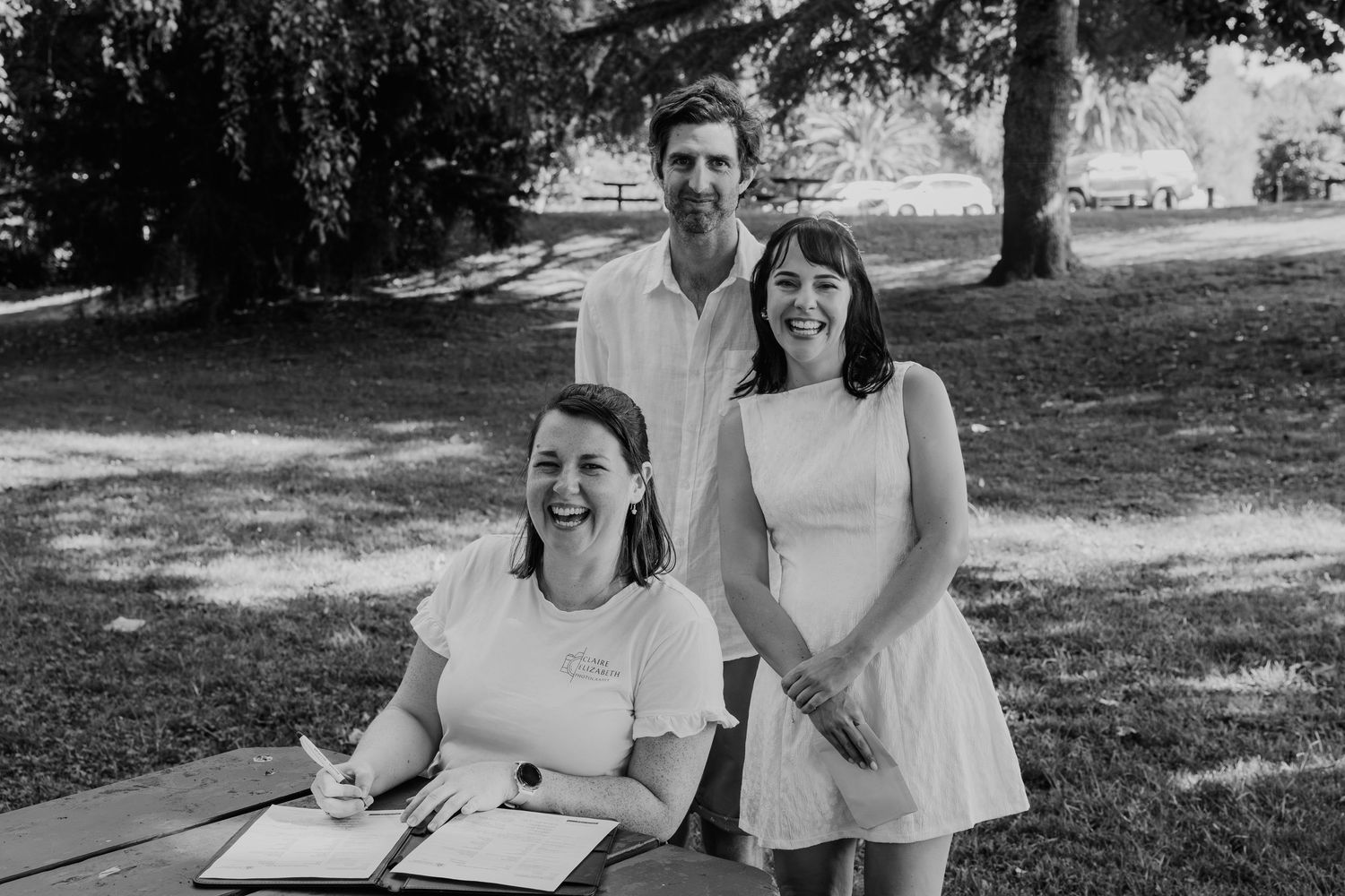 Three people smiling outdoors in a park, woman signing documents at picnic table, black and white photo.