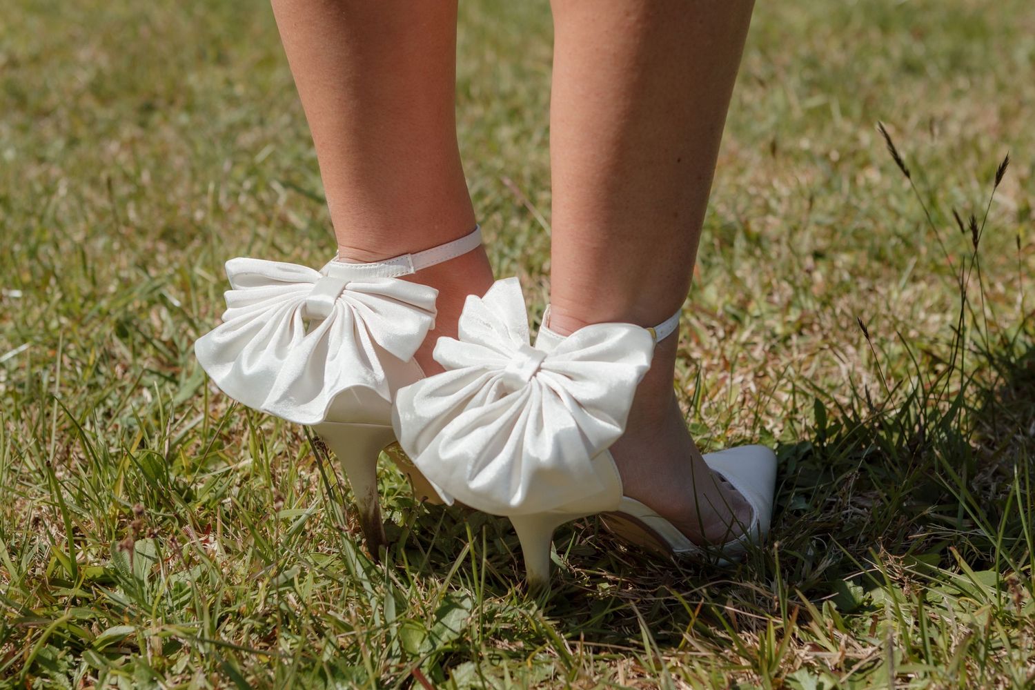 White bridal shoes with large satin bows on the ankle straps stand on grass in sunlight.