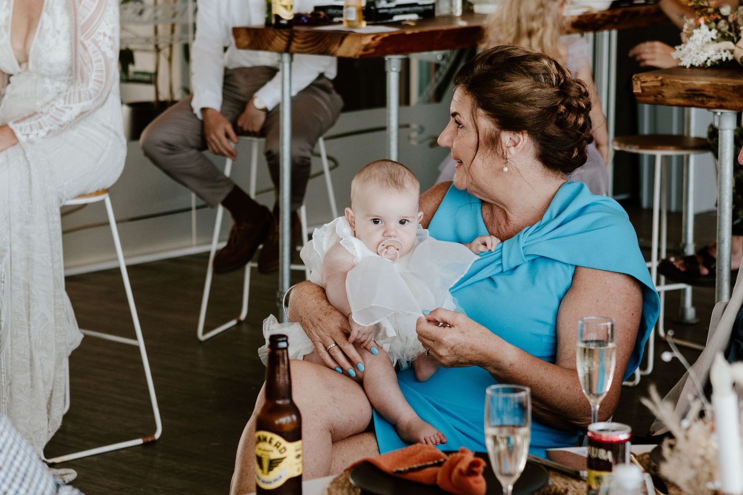 A baby in a white dress sits with someone in a blue top at a reception table with champagne glasses.