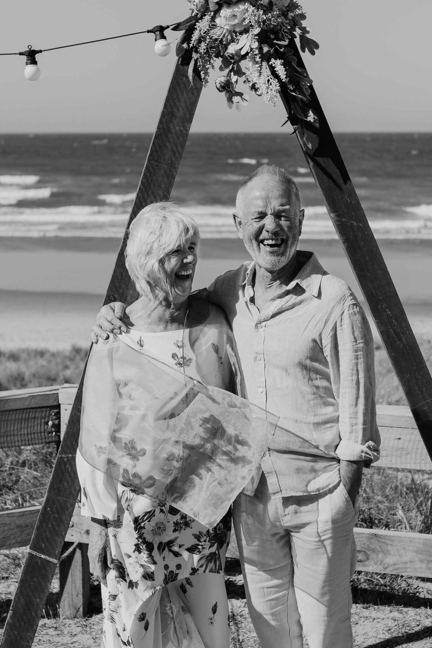 A couple shares a happy moment together under a decorated wooden arch by the beach on a sunny day.
