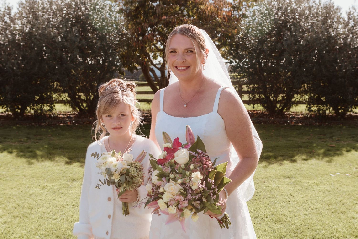 Two people in white dresses stand outdoors holding floral bouquets on a sunny day.