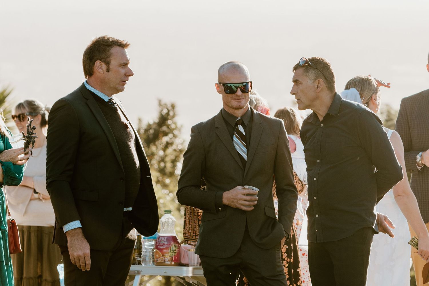Wedding party in black suits and dresses gather together at sunset during outdoor celebration.