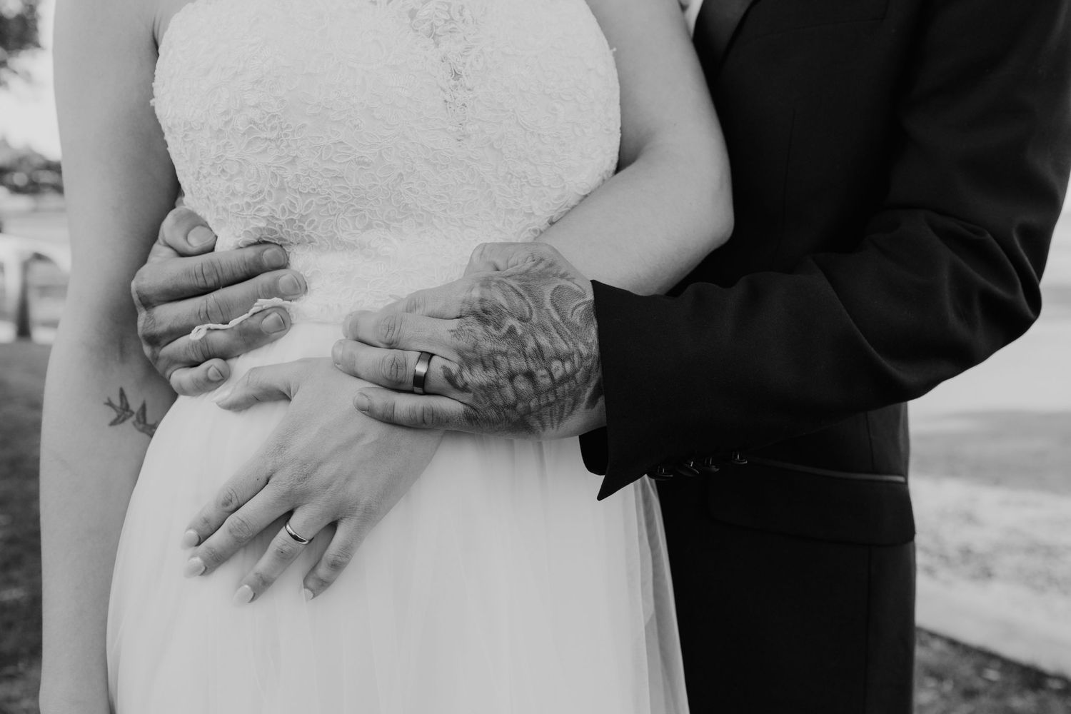 A black and white close-up of hands embracing during a wedding, showing rings and lace details.
