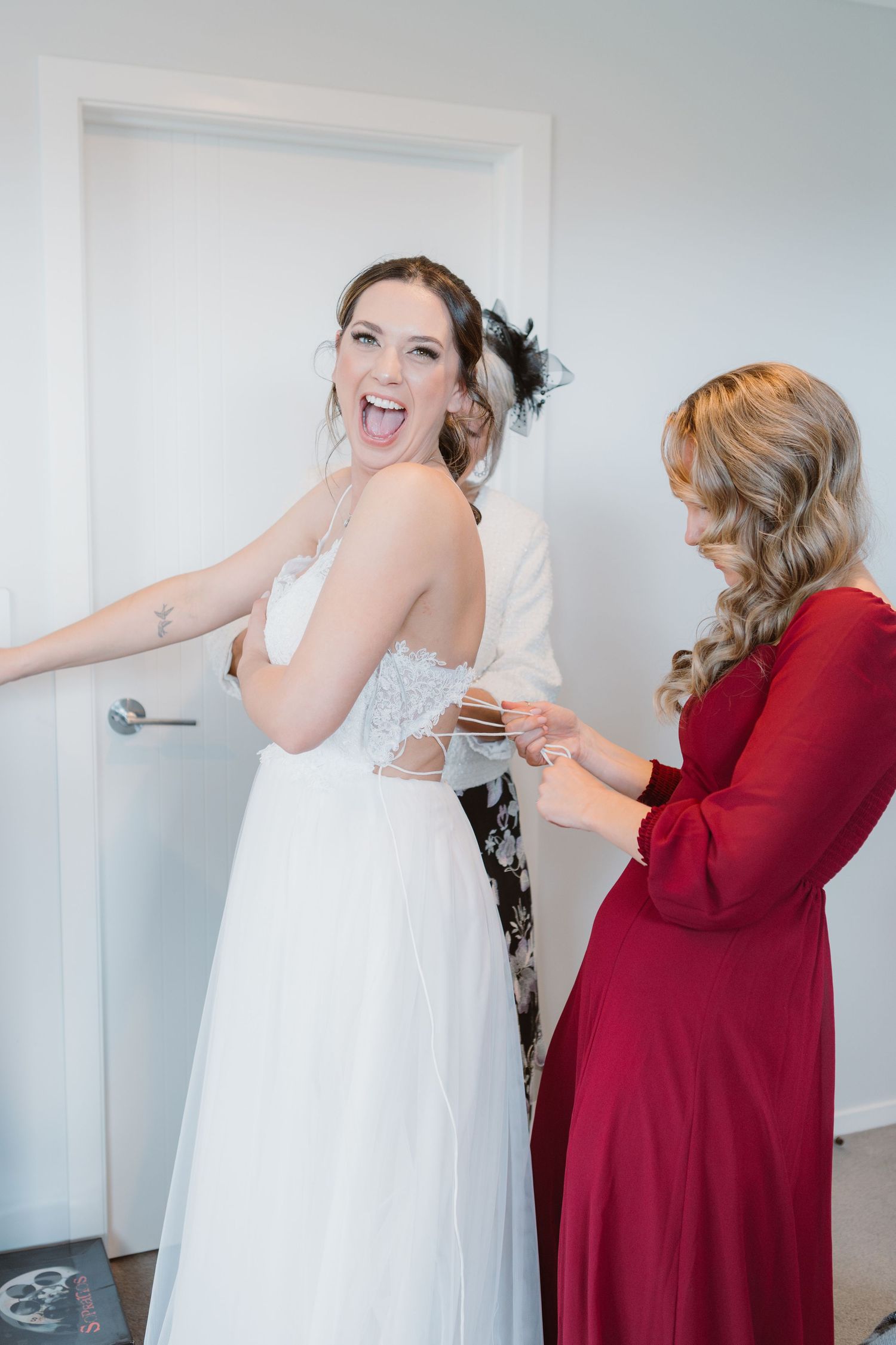 Wedding dress fitting moment with bridesmaid in burgundy dress helping with gown adjustments.