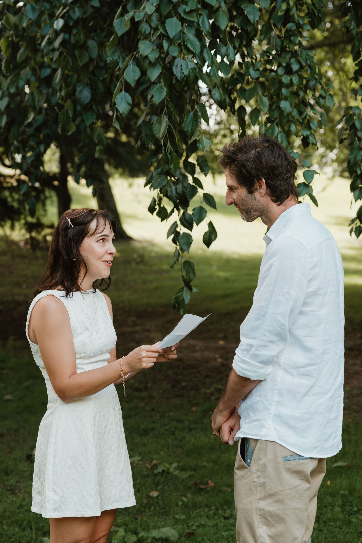Couple exchanging vows outdoors under a leafy tree, woman in white dress holding paper, man in linen shirt smiling.