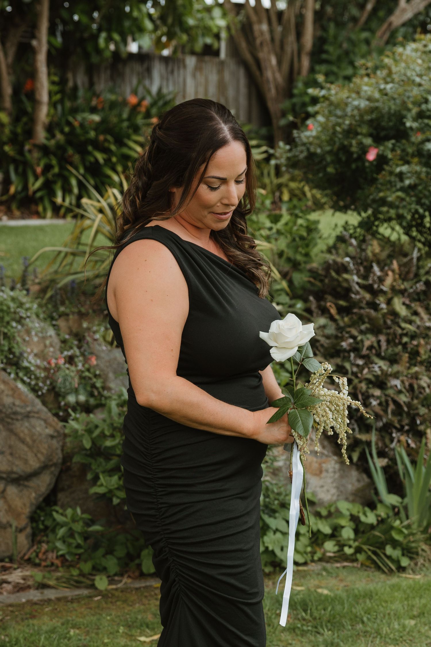 A woman in a black formal dress holds a single white rose while standing in a lush garden setting.