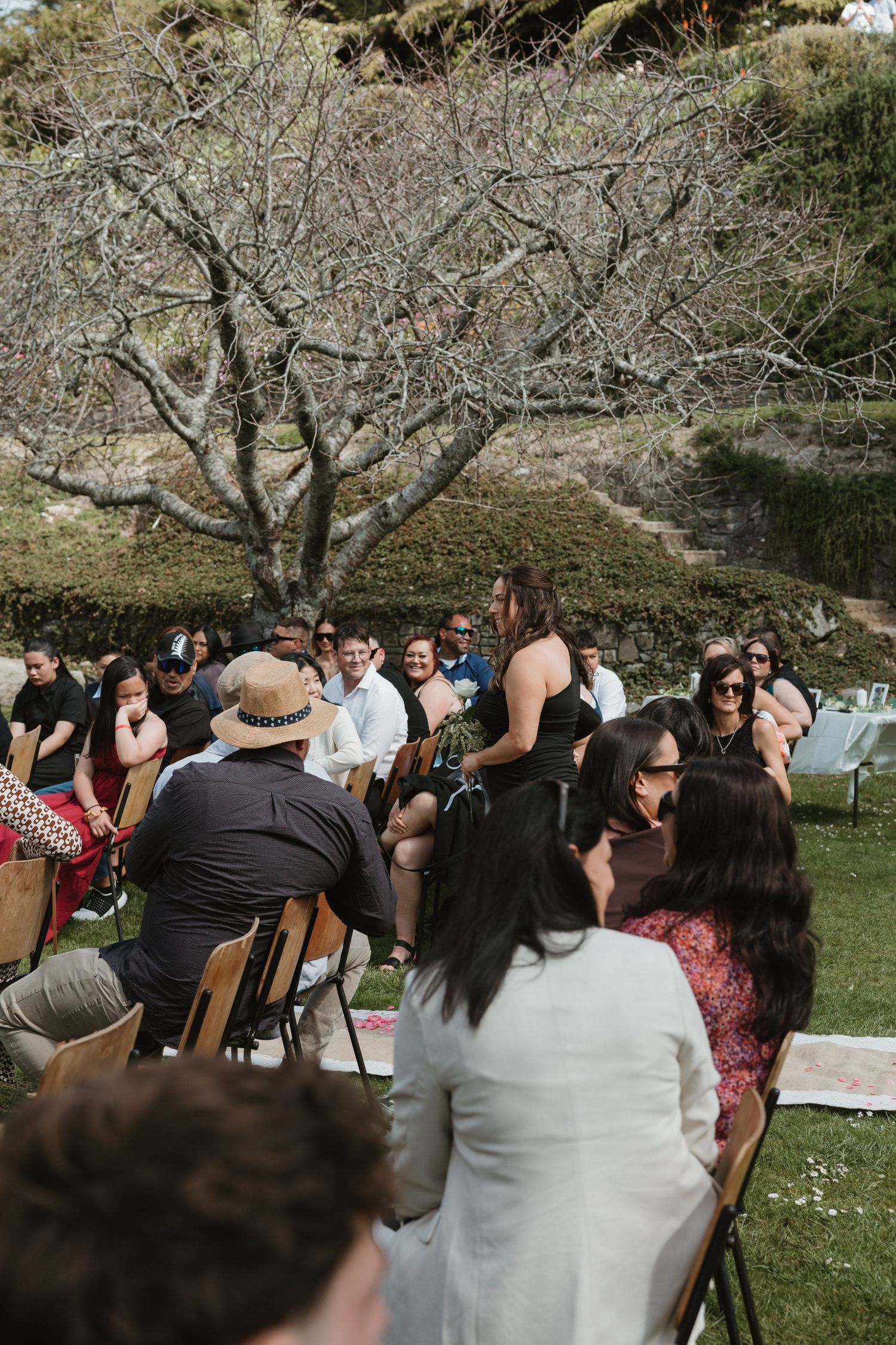 Outdoor gathering with people seated on chairs under a bare branched tree in a garden setting.