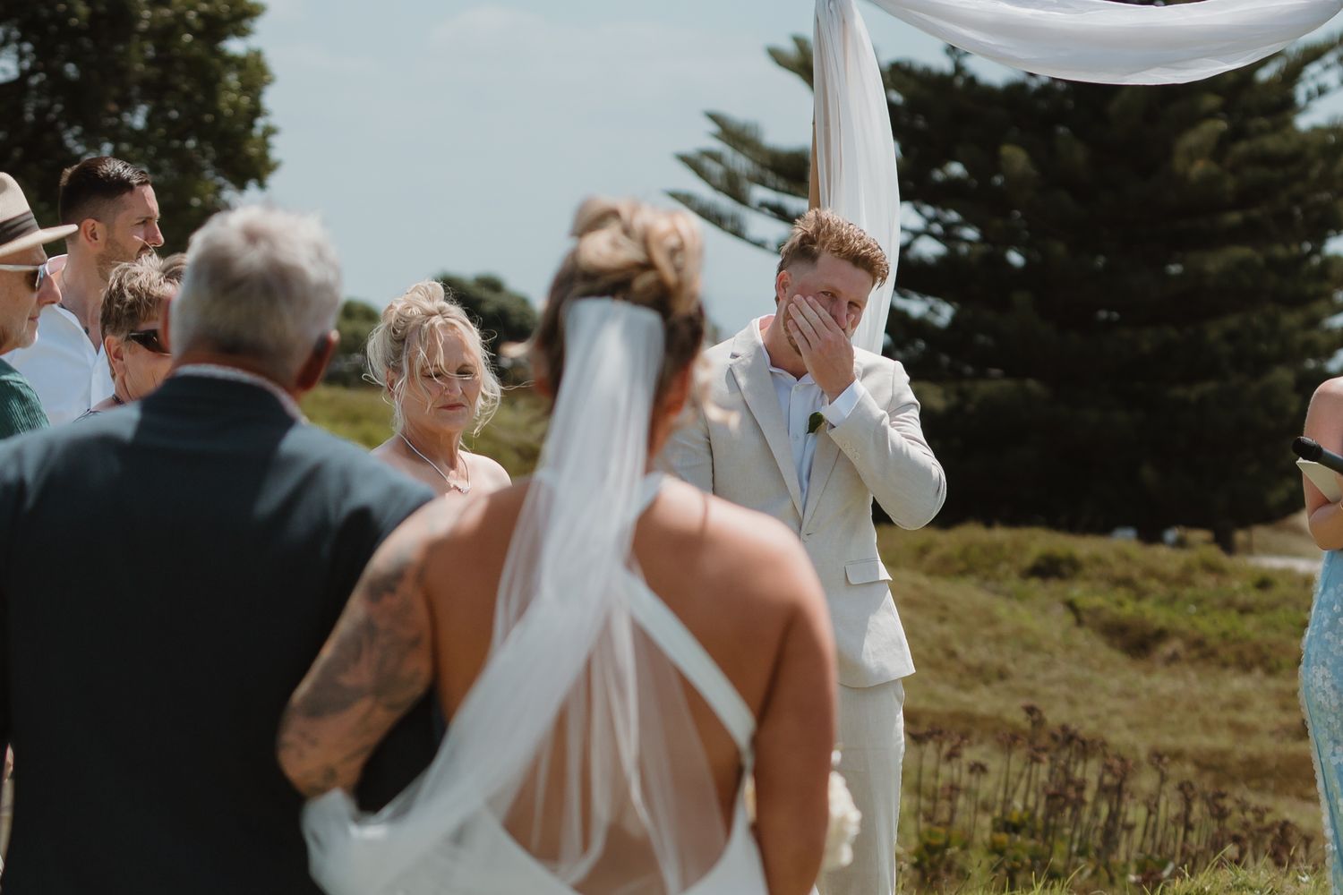 Emotional groom covers his face as bride walks down the aisle at outdoor wedding ceremony.