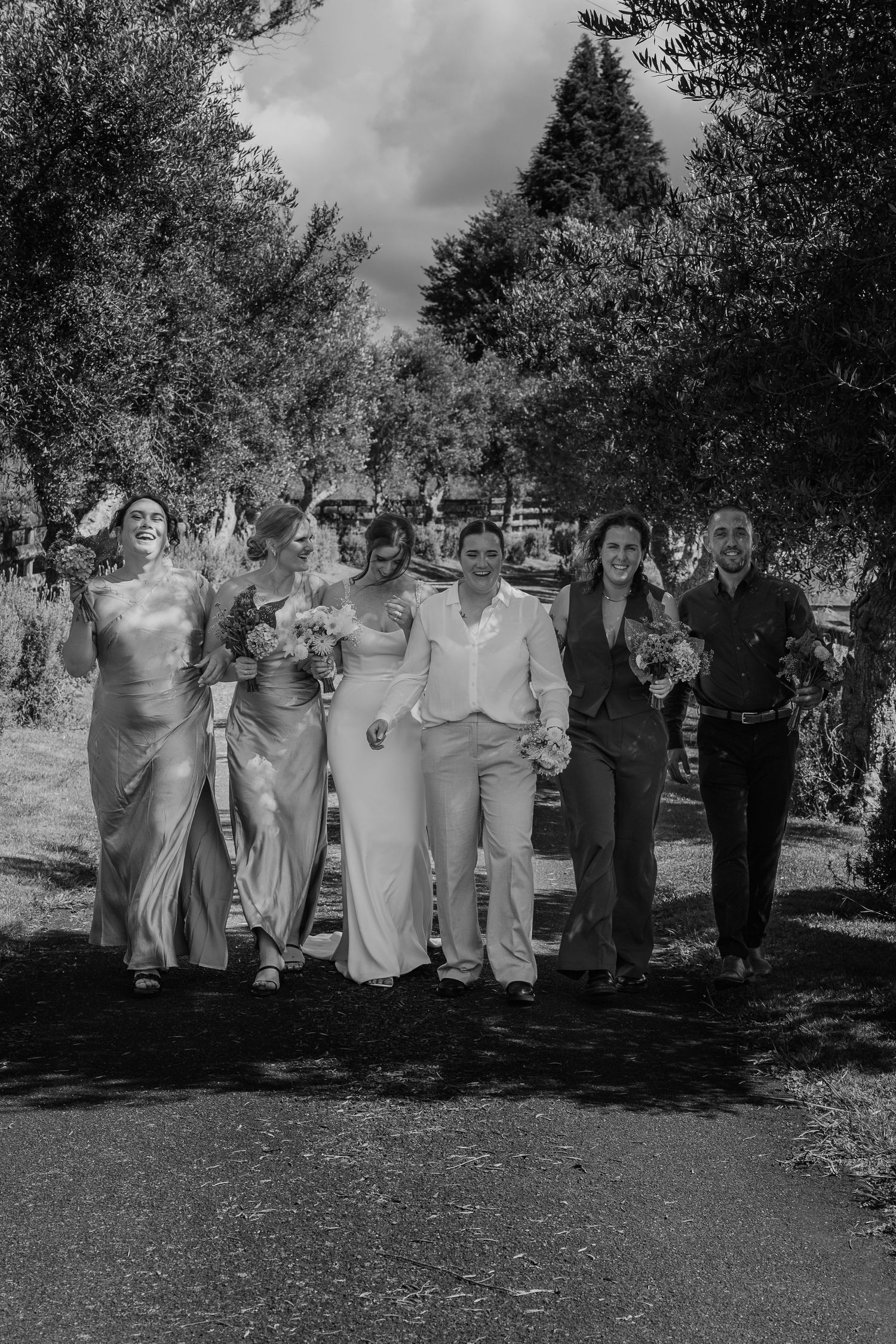 Black and white photo of a wedding party walking together outdoors, brides in white suits with bridesmaids and groomsmen.