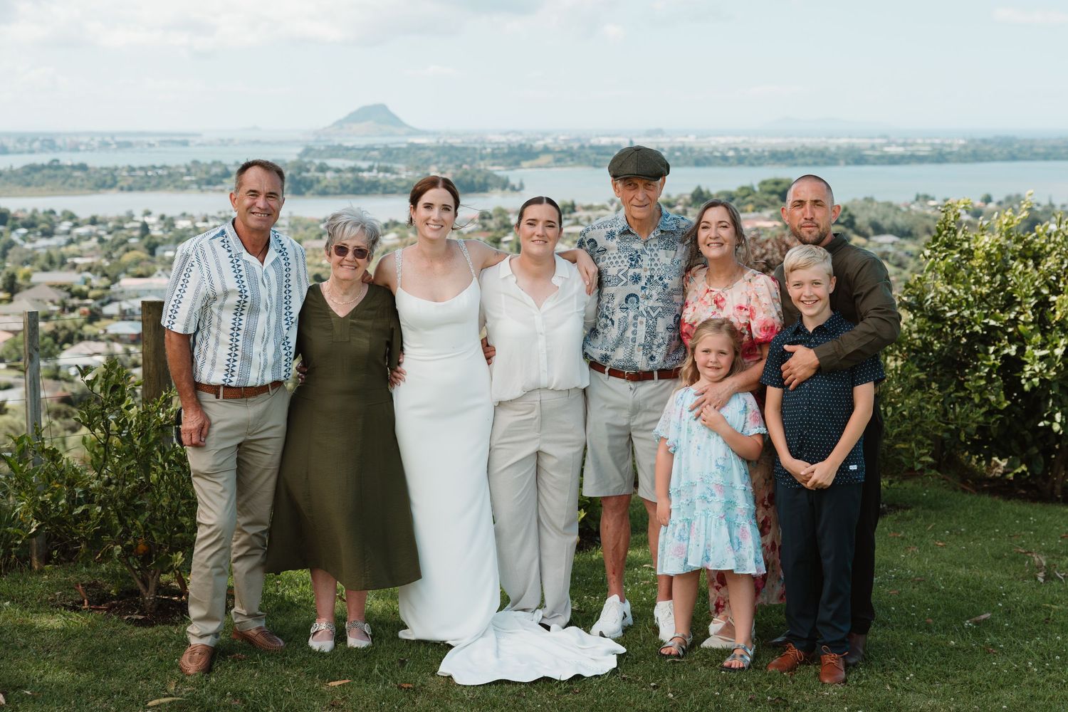 Wedding group photo with bride in white gown, family members smiling outdoors with scenic lake and town views behind them.