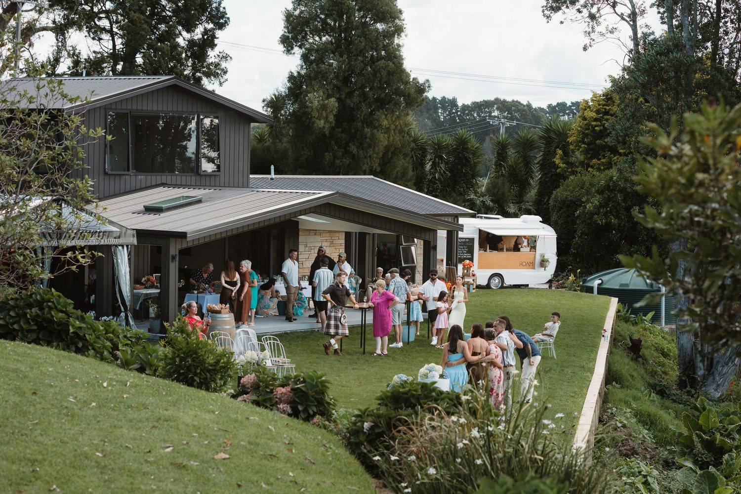 Guests gather on a lush green hillside lawn outside a modern home with a food truck during an outdoor party.