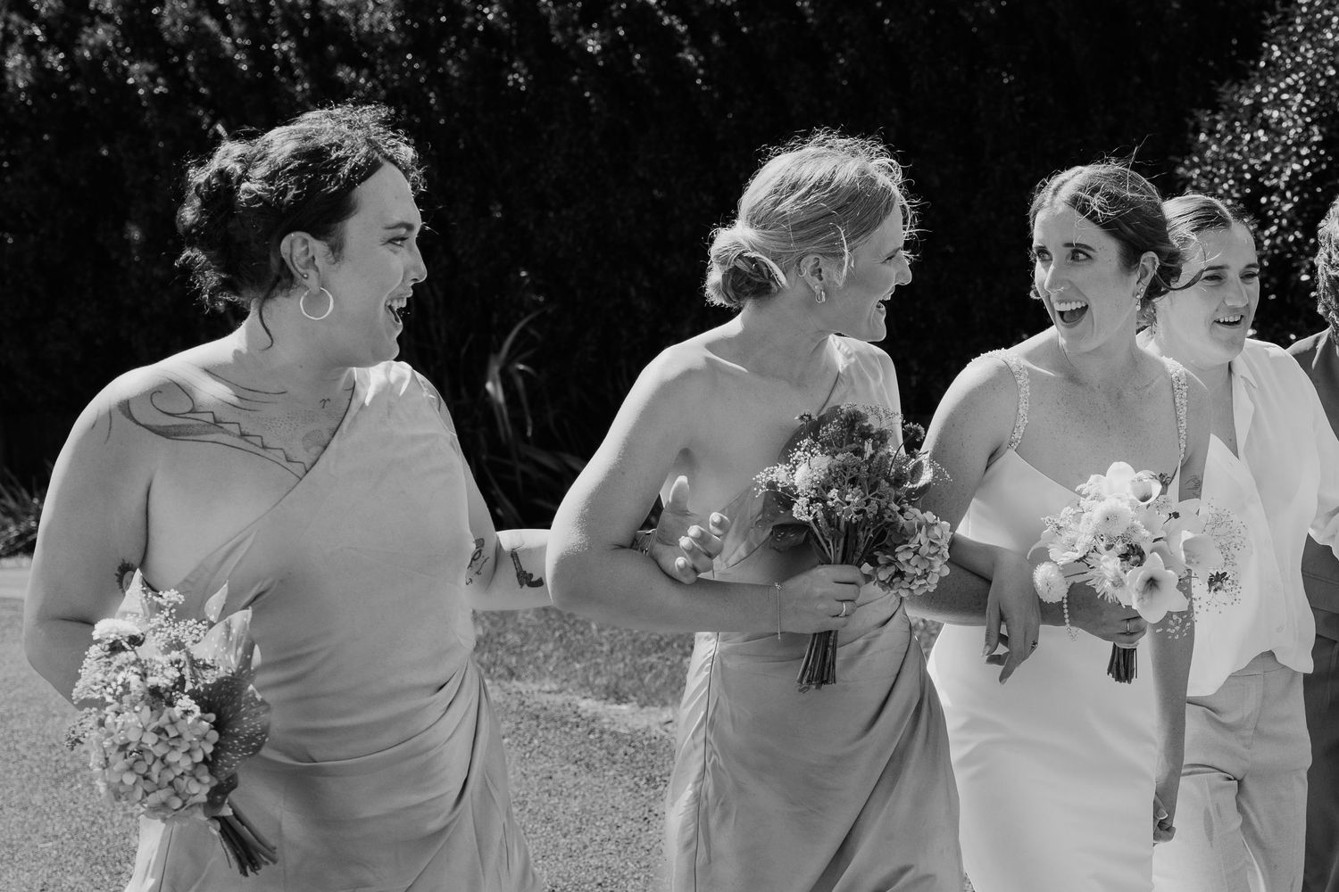 Black and white photo of bridesmaids walking together laughing, holding bouquets at an outdoor wedding.