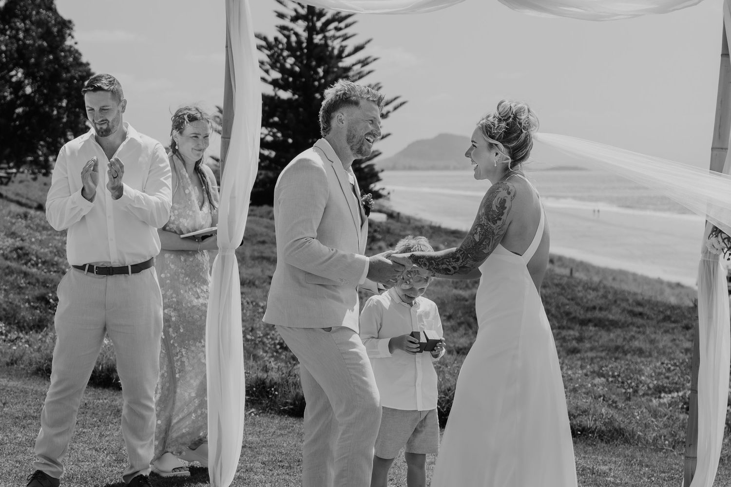 Black and white photo of a joyful outdoor wedding ceremony with couple exchanging vows under a draped arch.