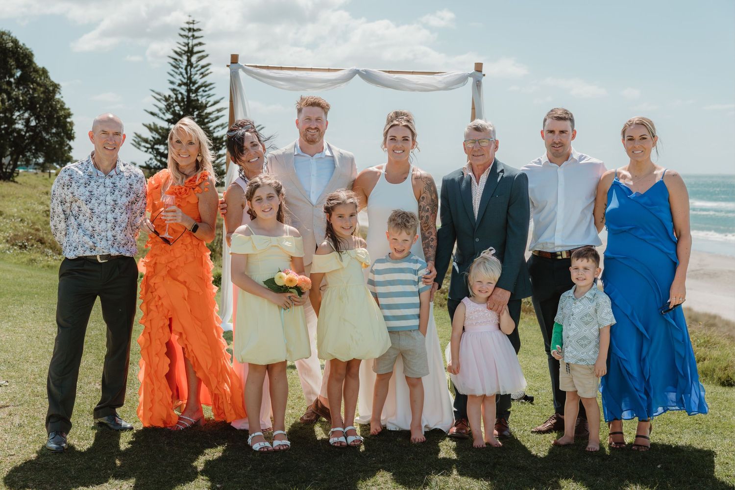 Wedding party posing outdoors near ocean with arch, bride in white, guests in colorful attire, children in foreground.