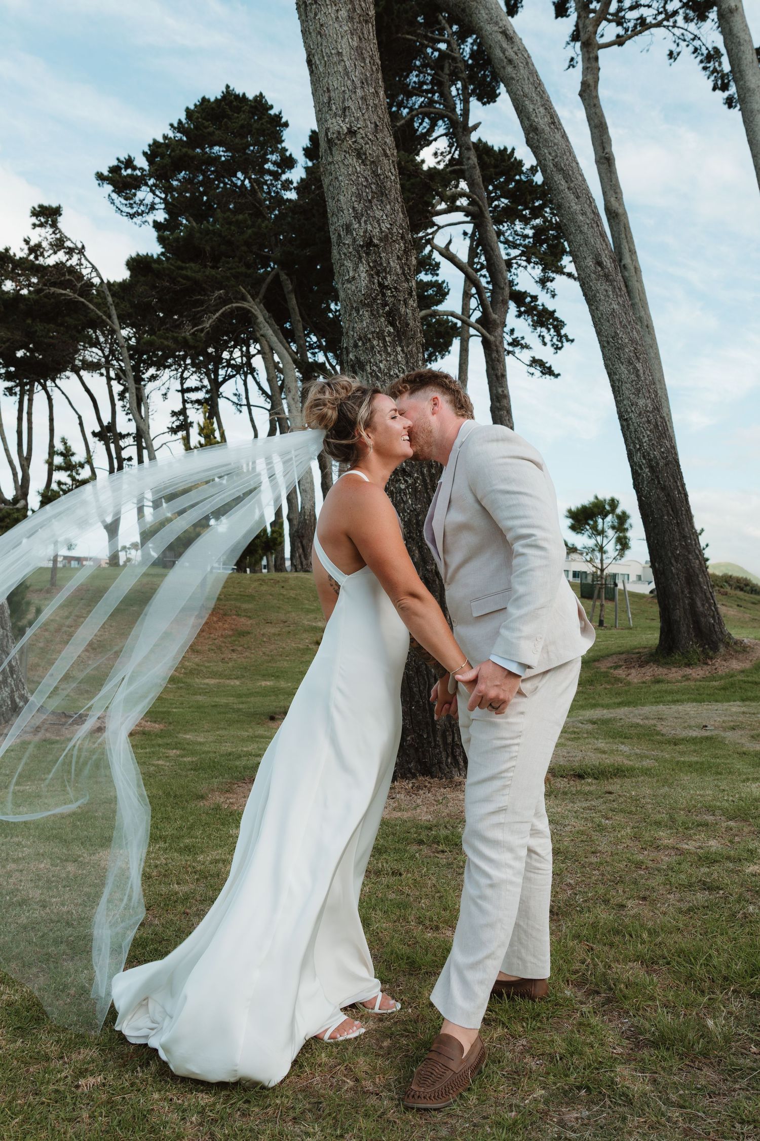 Bride and groom kissing outdoors near trees, bride wearing white gown with flowing veil, groom in beige suit.